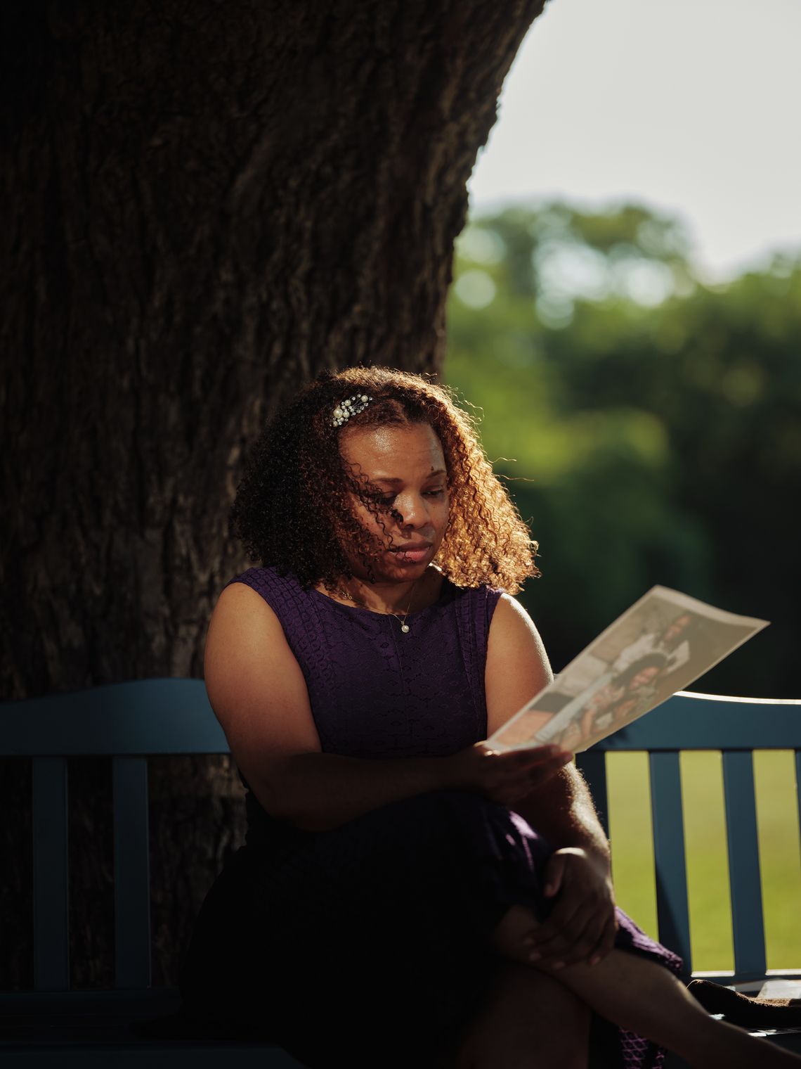 A Black woman wearing a purple dress, sits on a park bench looking pensively at a picture. 