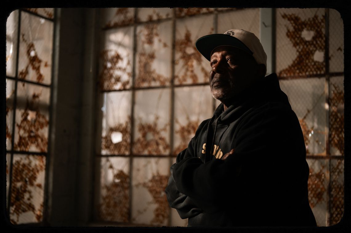 A photo shows Dorsey Nunn, a Black man with a white beard wearing a baseball cap and a hooded sweatshirt, standing in a space with cracked windows around him. 