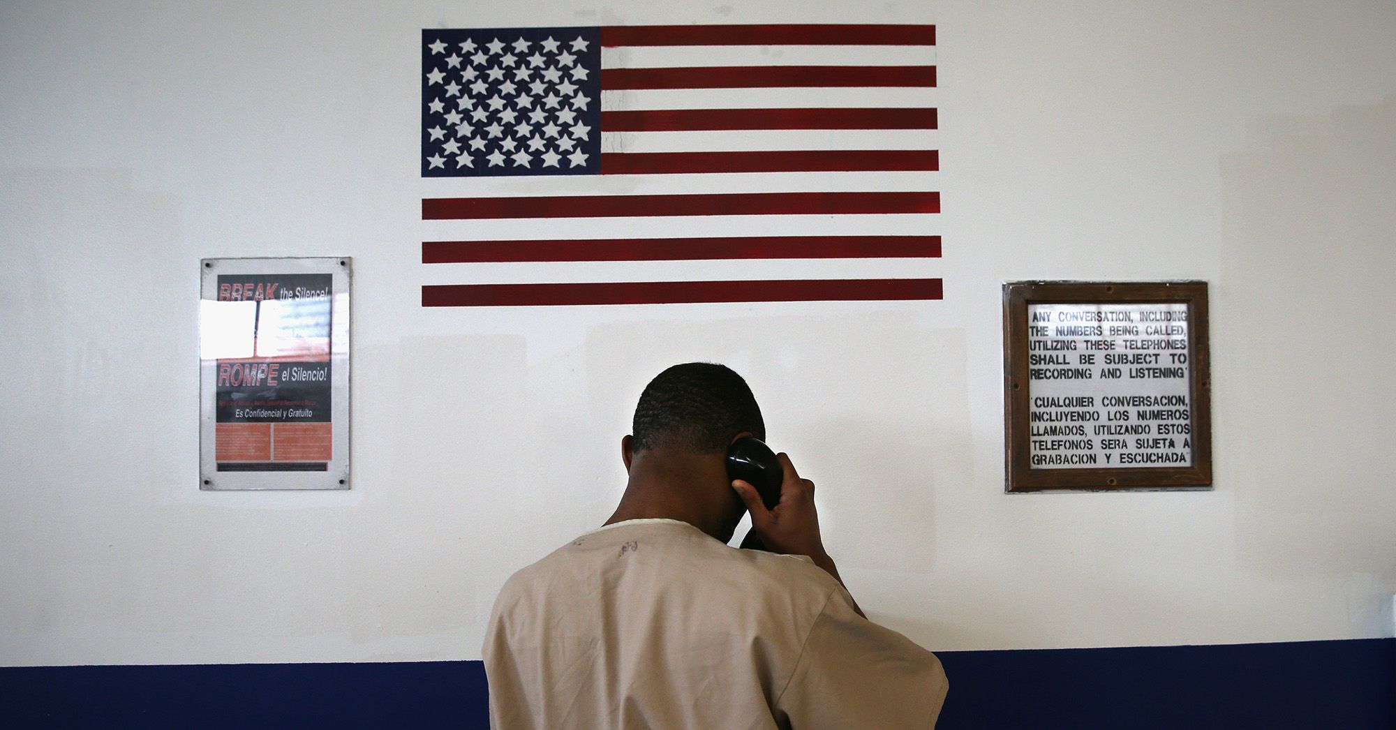 A Black man in a tan prison uniform stands in front of a black and white wall, holding a phone to his ear. A U.S. flag and two signs are on the wall. One sign reads, "Break the silence!" in English and Spanish. The second sign details that calls will subject to recording and listening.