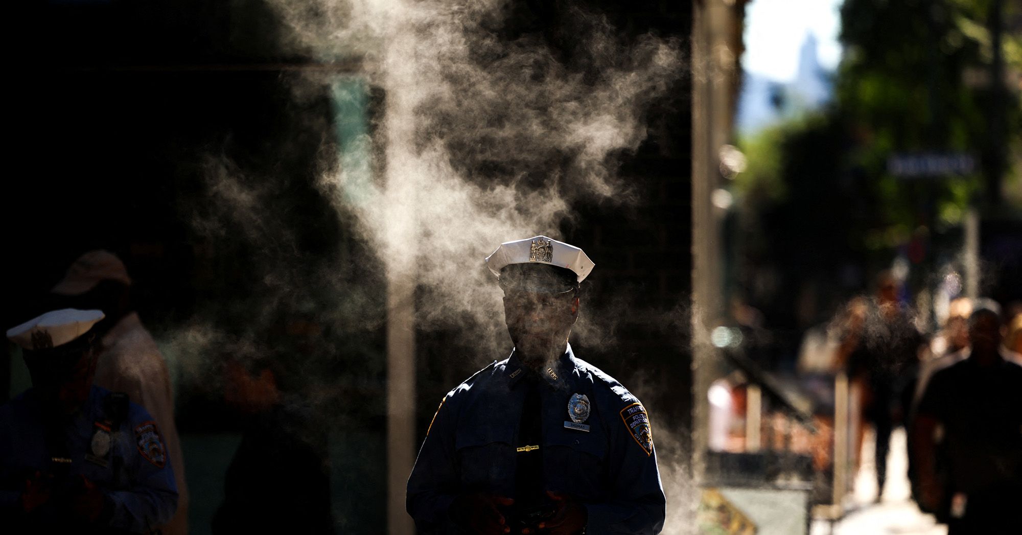 A police officer walks through a small cloud of smoke on a street. 