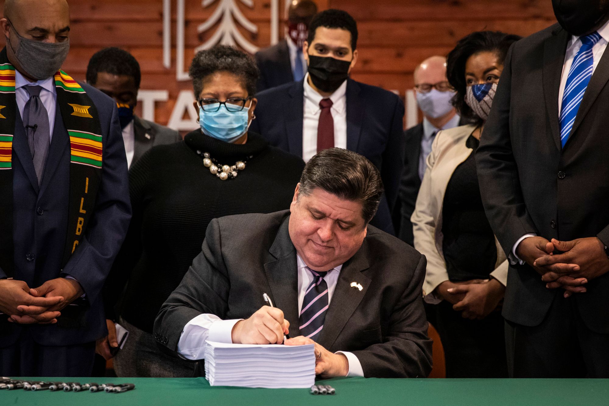 Gov. J.B. Pritzker, a seated man with light-toned skin and short-cropped dark hair, signs a bill that is composed of a high stack of papers. Eight masked people with varying skin tones and genders stand behind him.