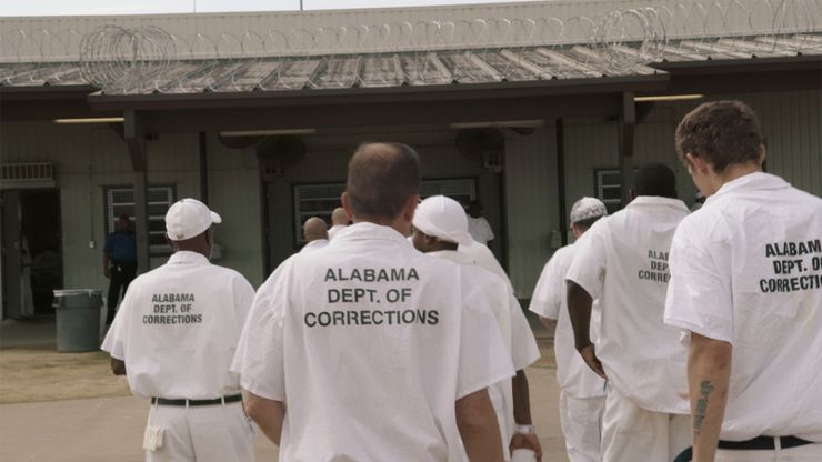 Several men wearing white prison uniforms with the words “Alabama Dept. of Corrections” printed on their backs walk toward a building. 