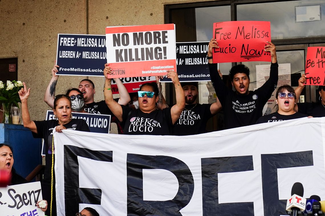 Outside of a building, a crowd representing mixed genders, ethnicities and ages stands holding signs above their heads with their mouths open. Some of the signs read “No more killing!” and “Free Melissa Lucio.” 