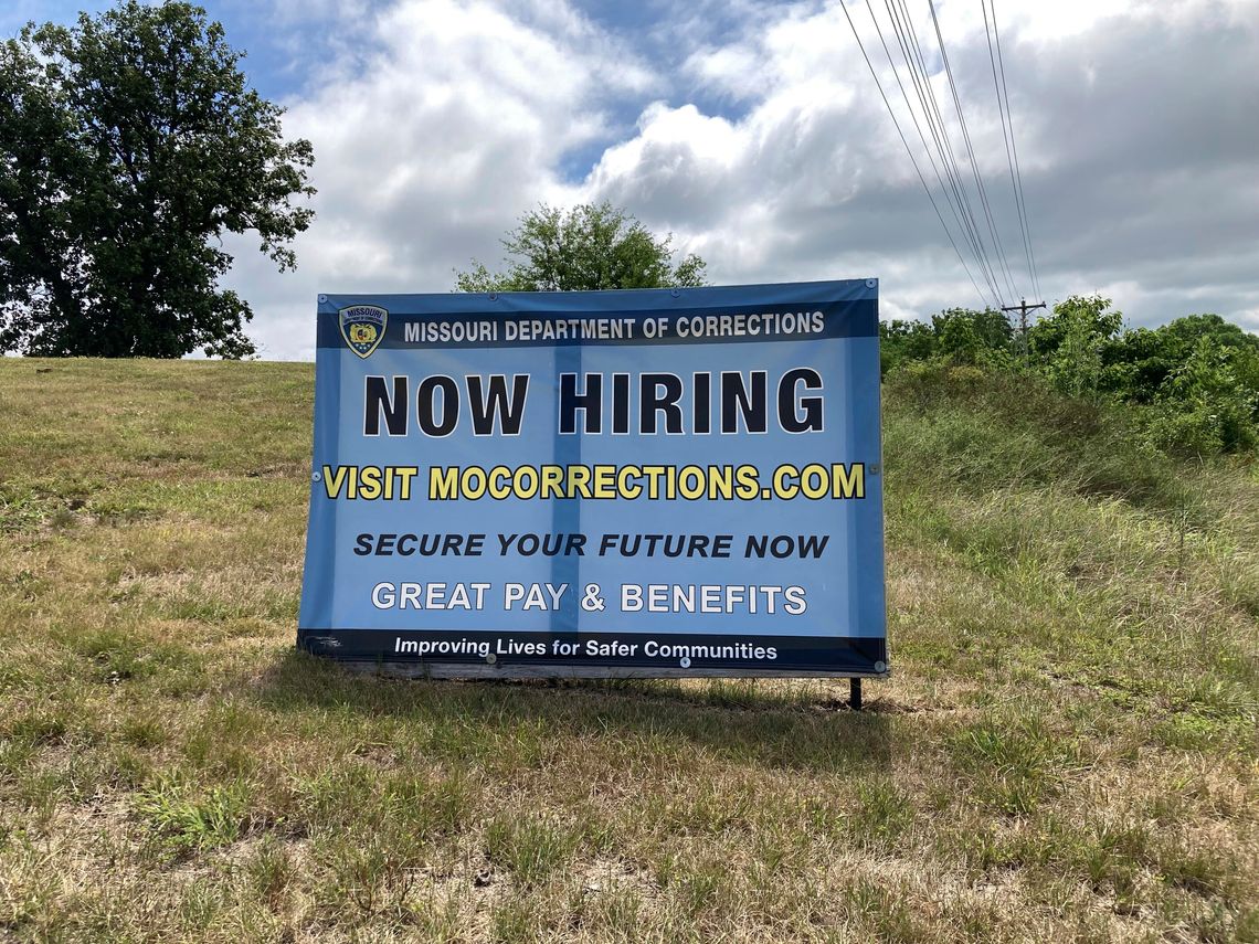 A large sign on a hillside from the Missouri Department of Corrections, reads “Now hiring. Secure your future now. Great pay and benefits.”