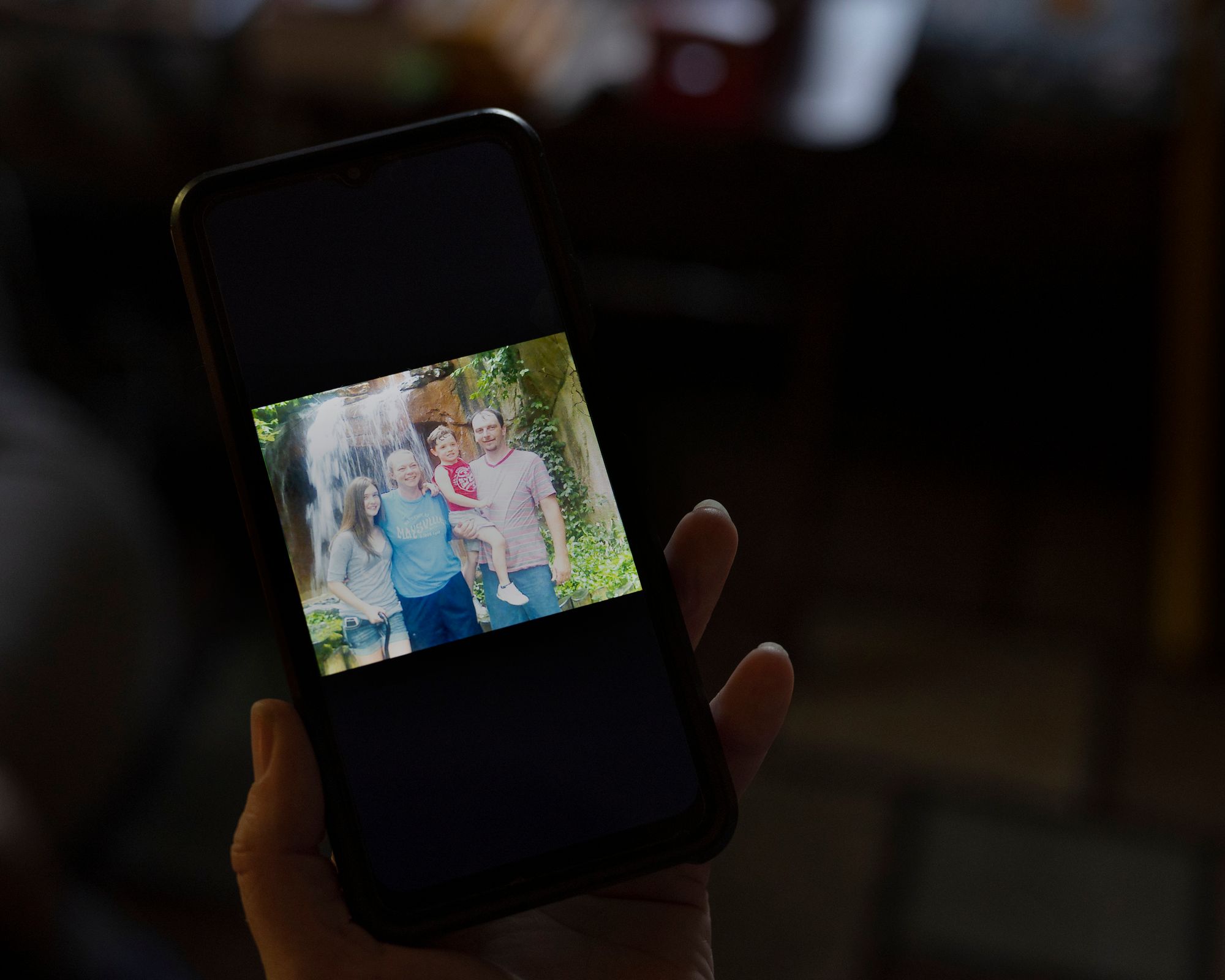 In a photo shown on a phone, Helphenstine, a White woman, stands in front of a waterfall next to her husband, a White man, while he holds their son.