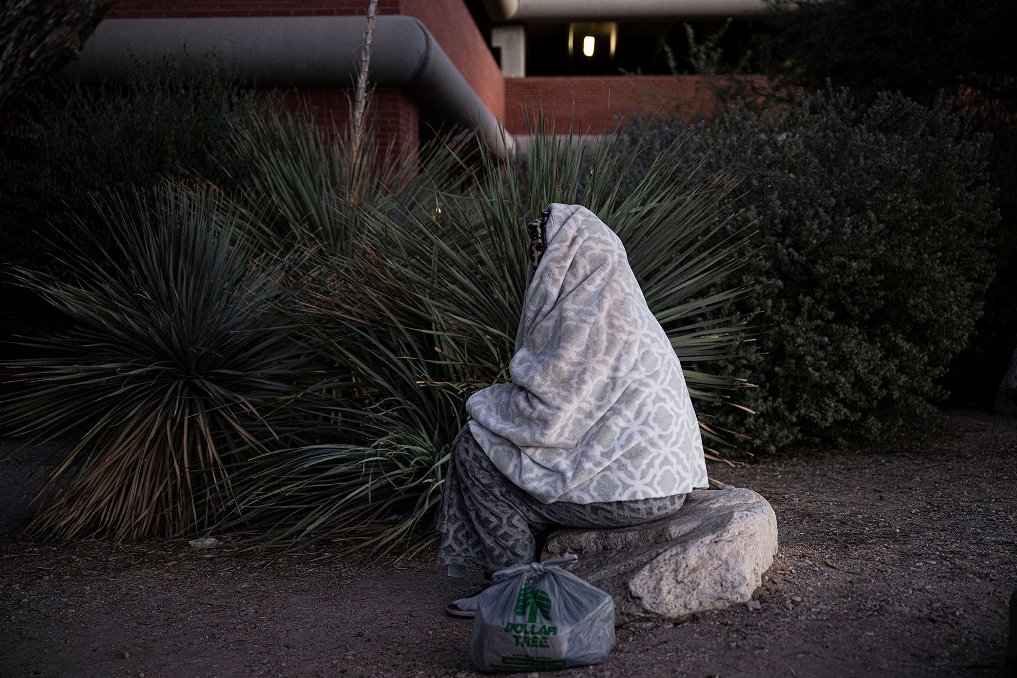 A photo shows a person, covered with a blanket, sitting on a rock outside. A Dollar Tree plastic bag is placed next to them. 
