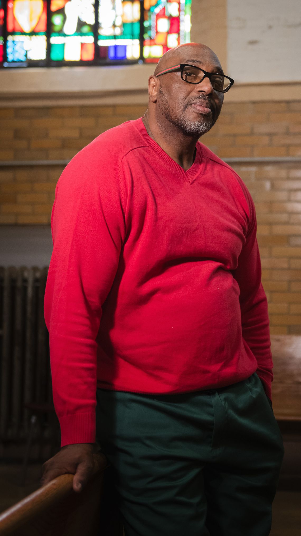 A bald, Black man wearing glasses and a red sweatshirt leans against the back of a church pew. Behind him on the wall is a multi-colored stained glass window. 