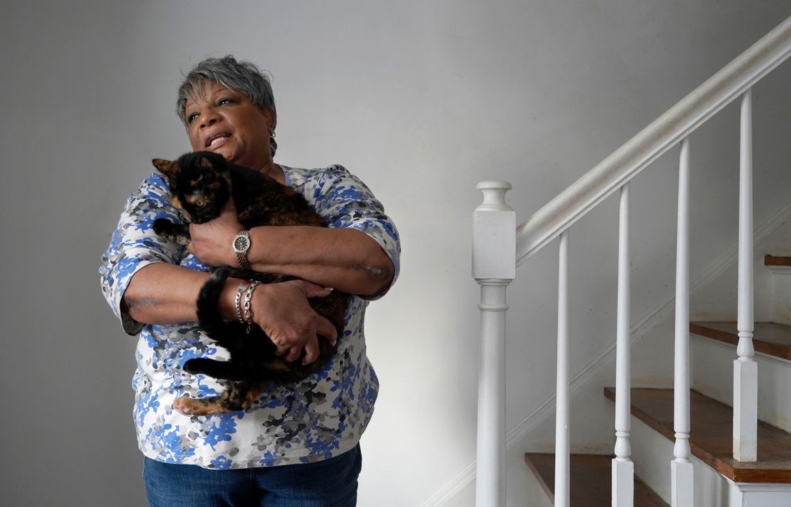 A photo of a Black woman in her 60s holding a cat next to a set of stairs. 