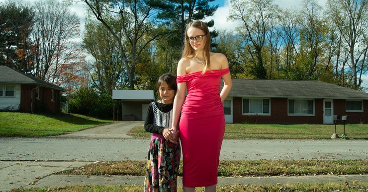 A White woman, wearing glasses and a red dress, holds the hands of her young daughter, who is wearing a black long-sleeved shirt and patterned dress. They stand in front of their house. 
