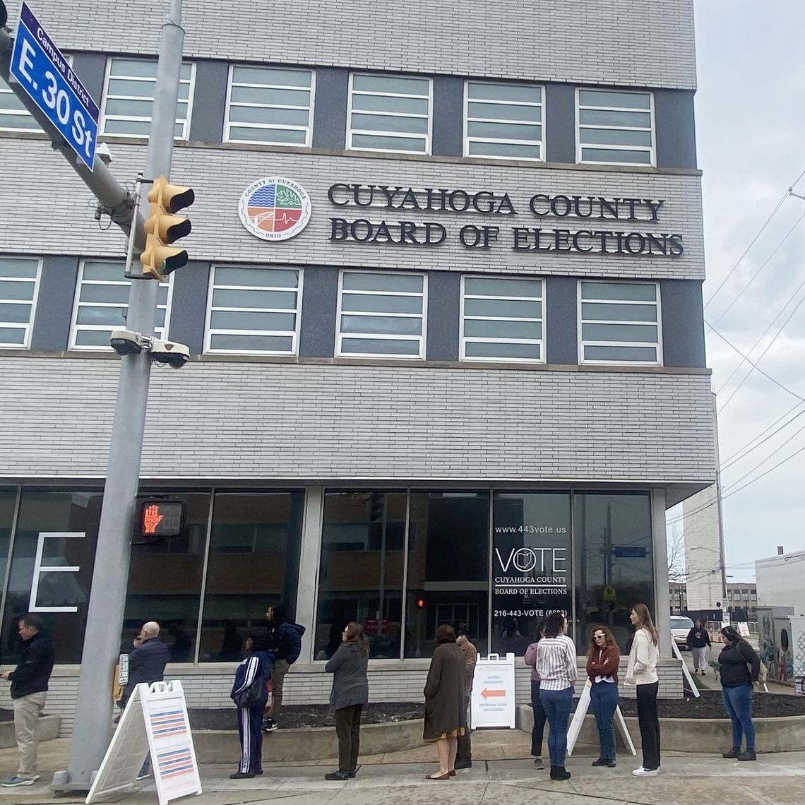 A photograph of more than 10 people lining up outside a gray building with a sign reading “Cuyahoga County Board of Elections.” 