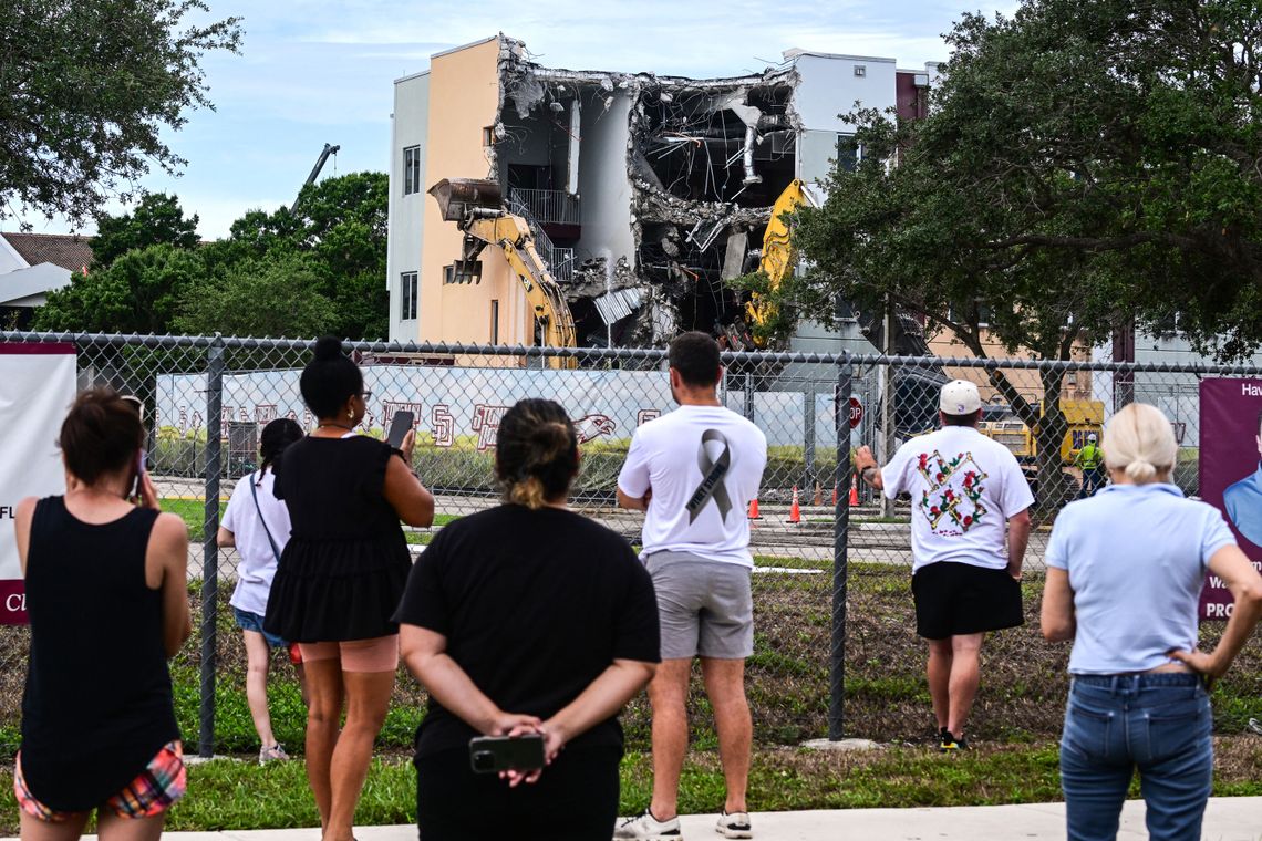 A photo of a group of people with their backs toward the camera watching a construction crew demolishing a school building behind a chain link fence. 