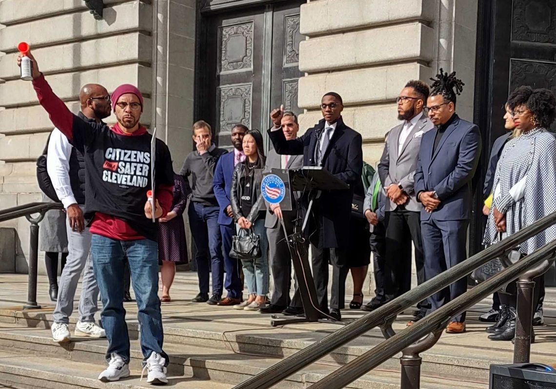Kareem Henton, a Black man, raises his arm while wearing a shirt that says “Citizens for a Safer Cleveland” on it, in front of a group of people surrounding Mayor Justin Bibb, a Black man, at a podium outdoors. 