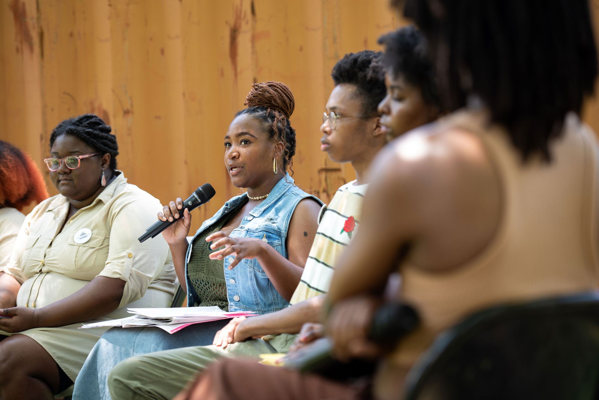 A photo of a Black woman with her hair in a bun speaking into a microphone while sitting on a panel.