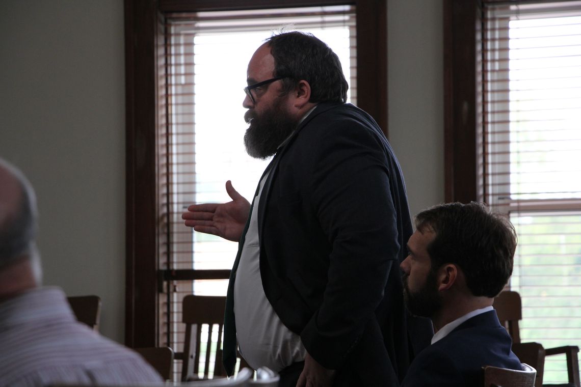 A photo shows a White male attorney, wearing glasses and a black suit, standing inside a courtroom. 