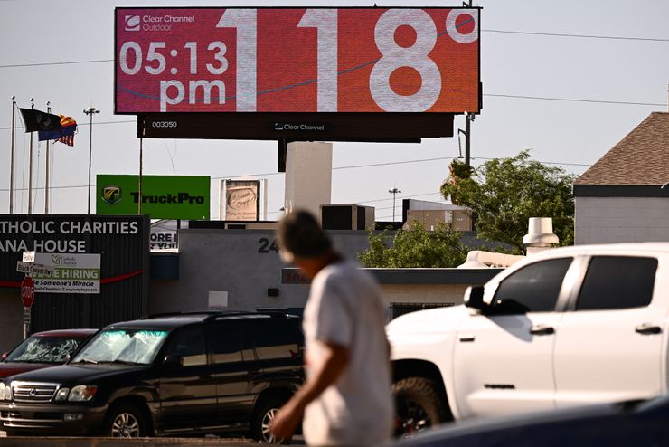 A billboard display reads "118 degrees, 5:13pm, Clear Channel Outdoor." A person walking through a parking lot is out of focus in the foreground.