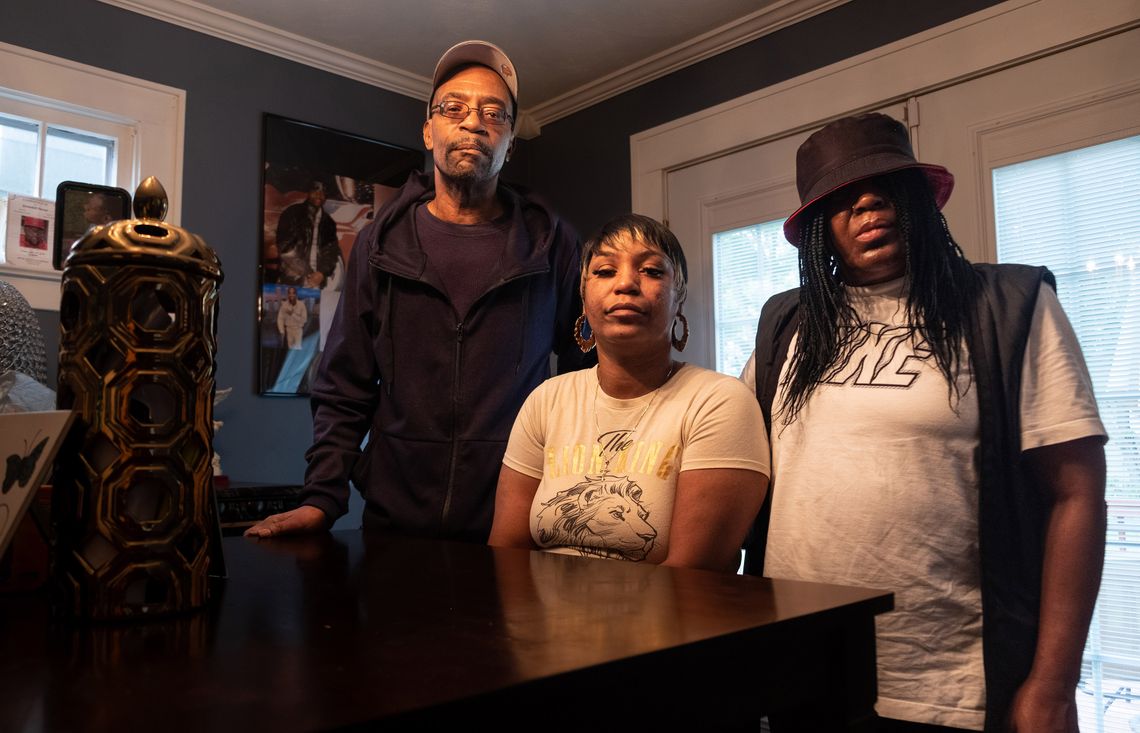 Bobby Everson is wearing a cap and navy sweatshirt. Ebony is wearing a white graphic T-shirt. Sabrina is wearing a black bucket hat, black vest and white T-shirt. The family members all have dark brown skin. They stand in a corner of their home.