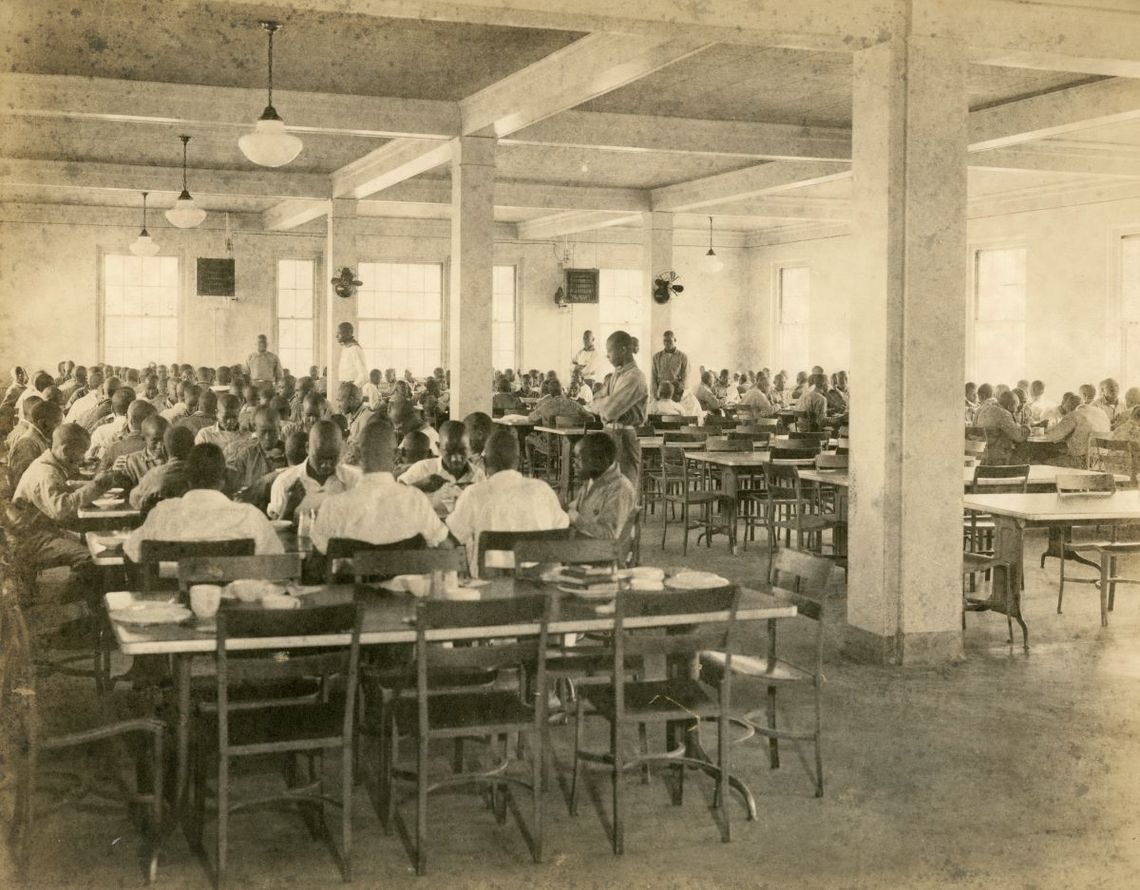Groups of Black boys, dressed in collared shirts, sit at about a dozen tables. There are some windows to the space, and pillars visible.