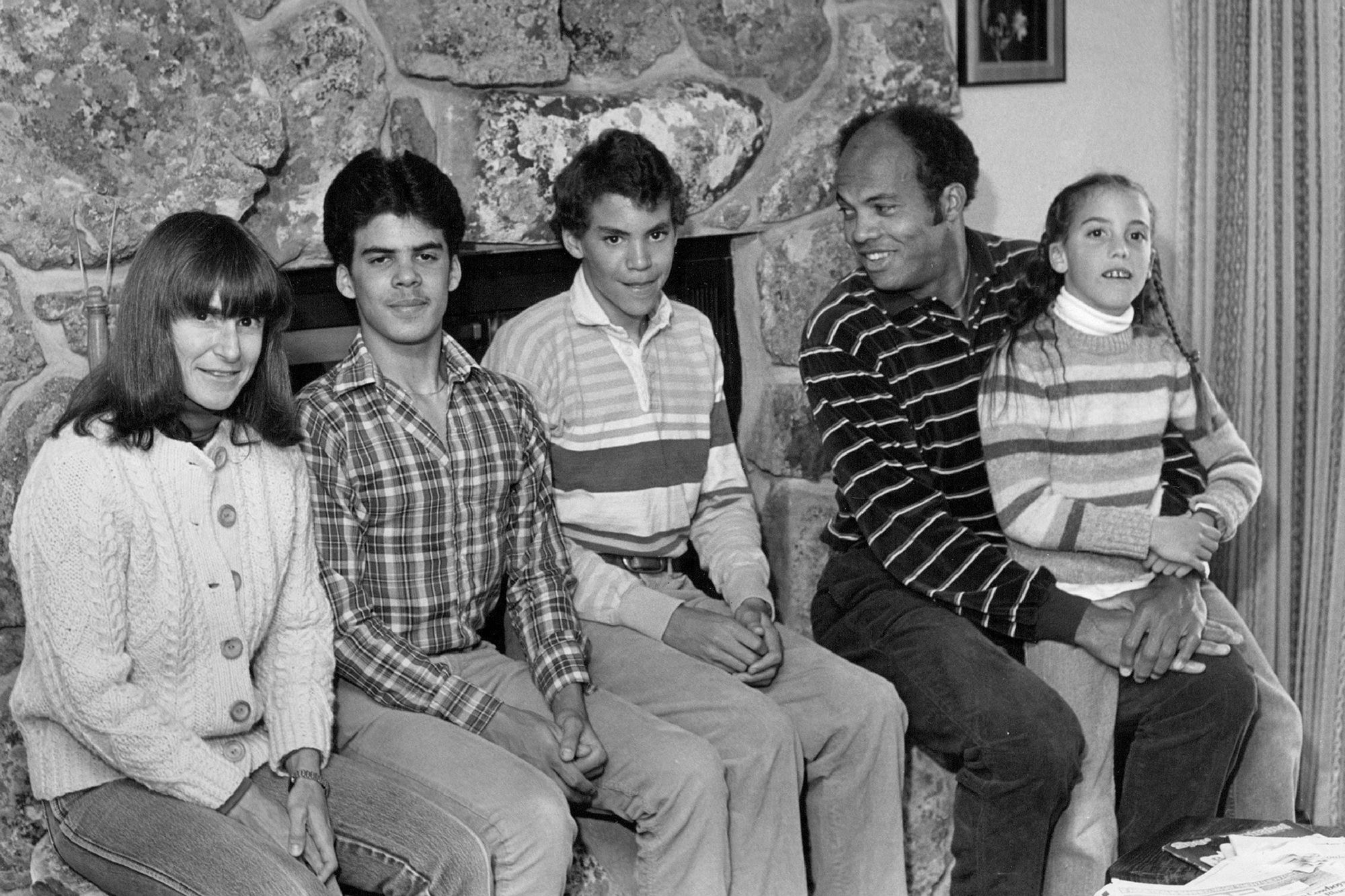 A black and white image shows a family of five sitting in front of a fireplace inside a home. 