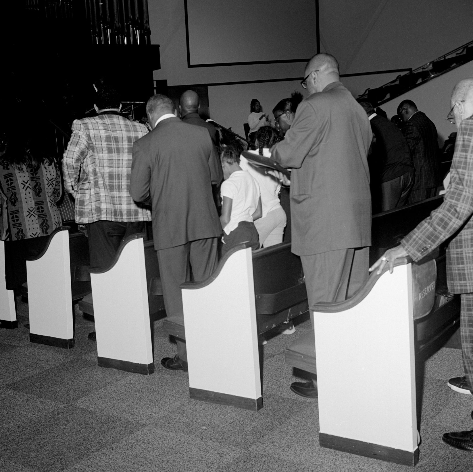 About a dozen people, formally dressed, stand among rows of church pews. 