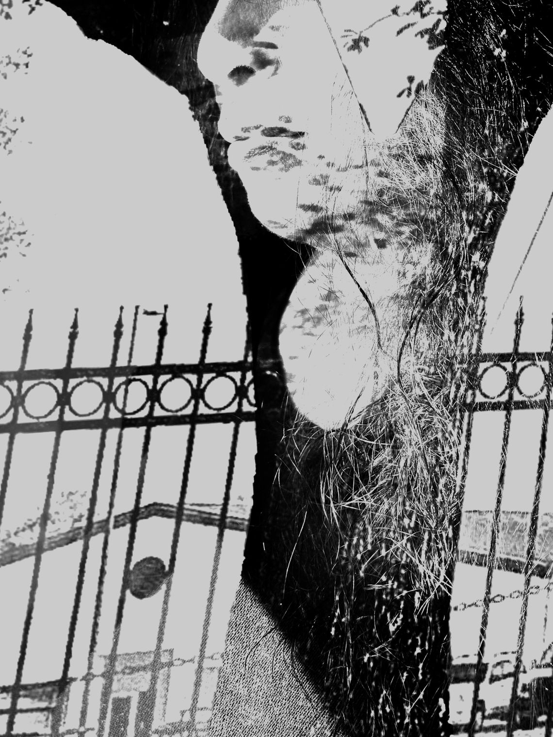 A black-and-white composite photo shows a woman whose nose and mouth are visible. Much of the photo is obscured by another photo layered on top in a double-exposure effect, which has a tree trunk, metal fence and a building on it.