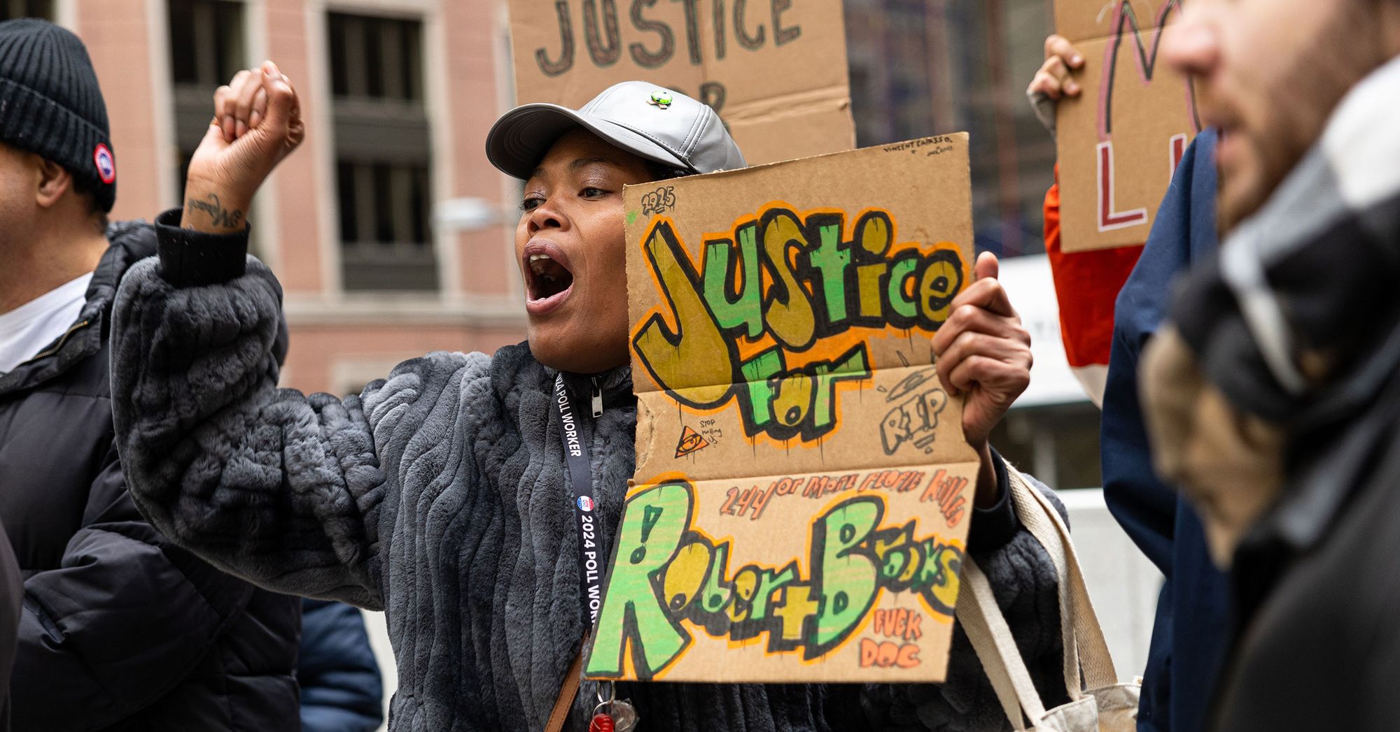 A Black Woman, wearing a silver cap and gray jacket, holds a sign that reads "Justice for Robert Brooks" during a protest. Protesters holding signs stand behind her.