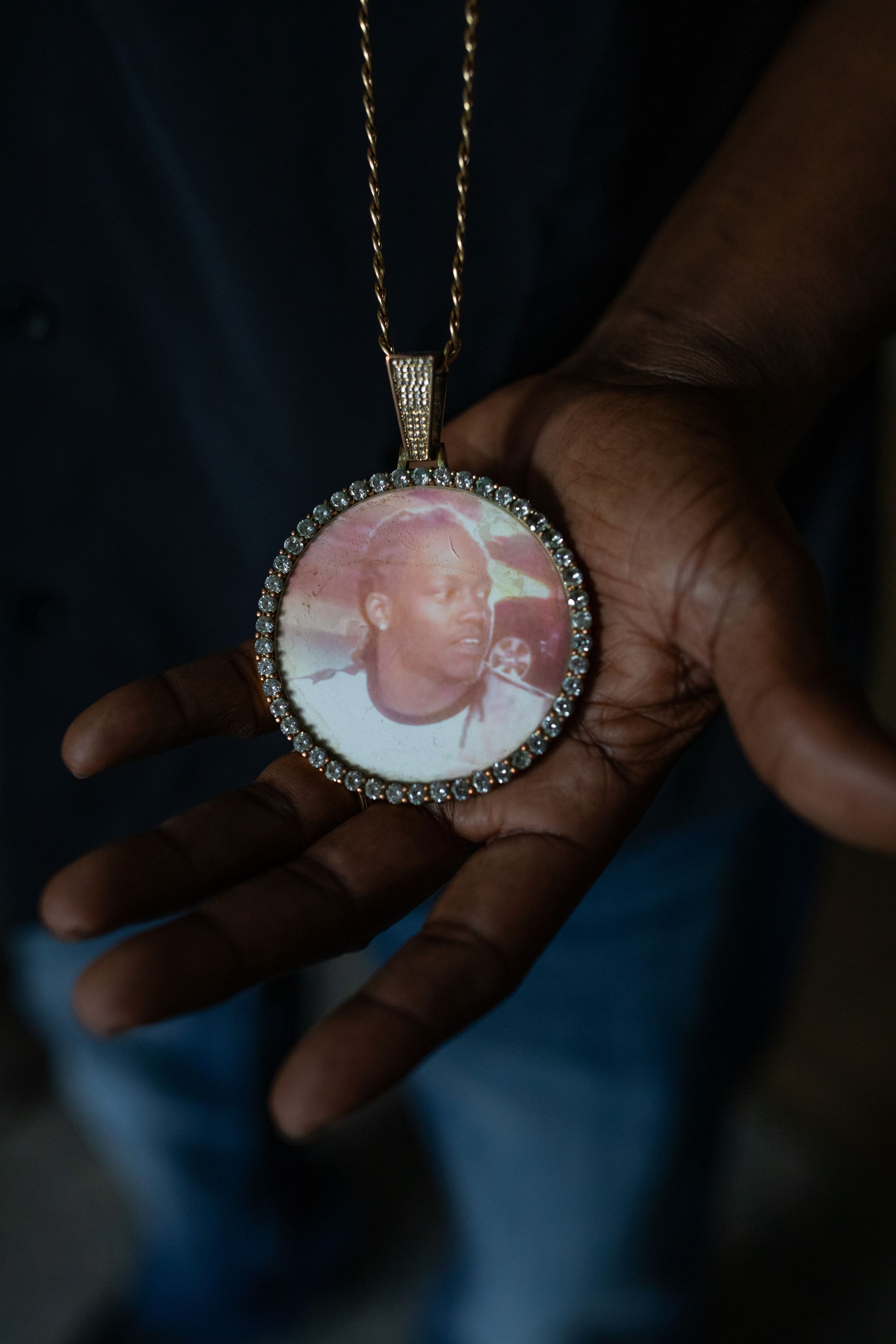 A photo shows a Black person’s hand holding a necklace with a large circular pendant with a picture of Brandon Webster, a Black man, on it. 