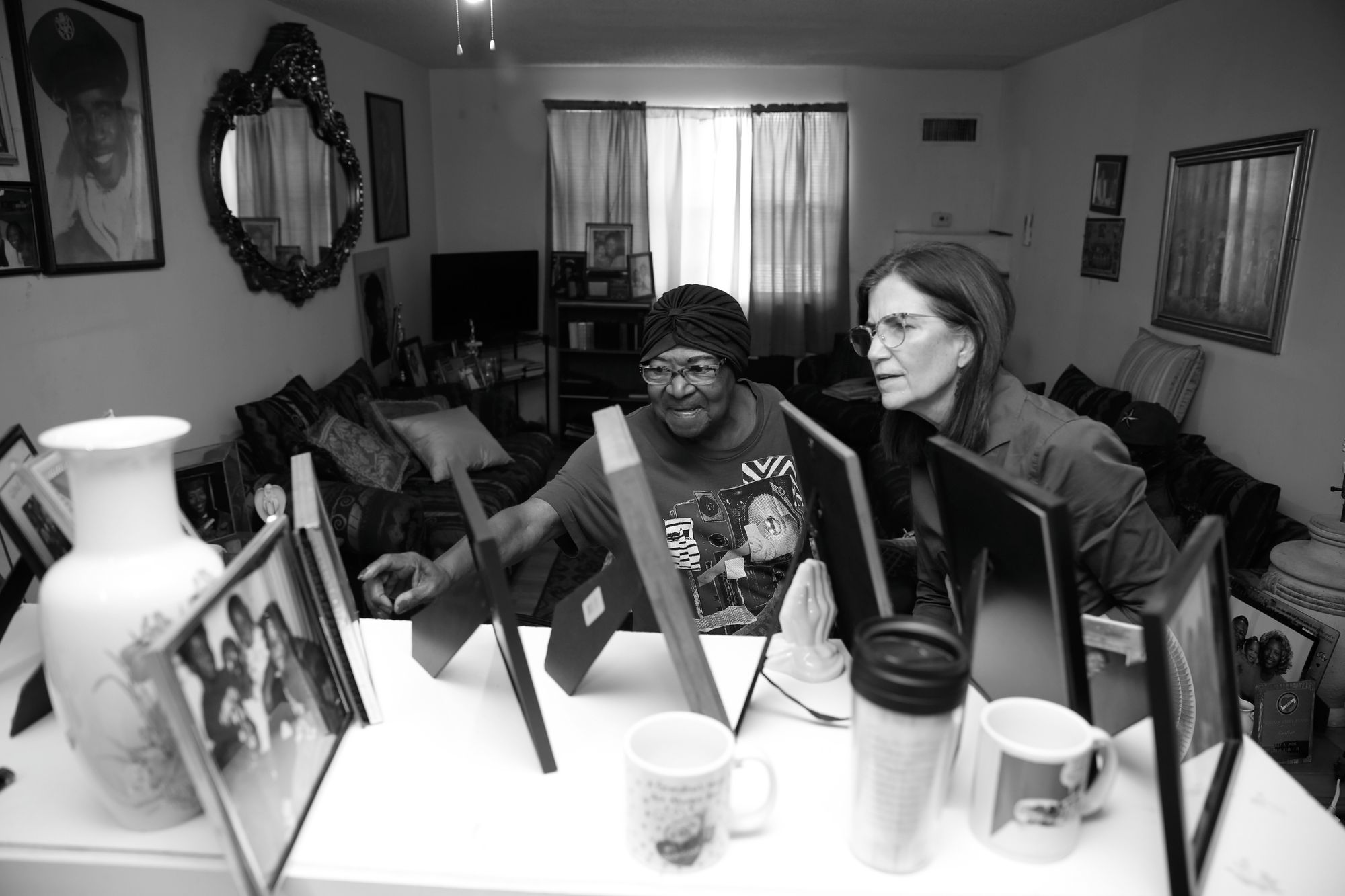 A Black woman points out a framed photograph in her living room to a White woman.