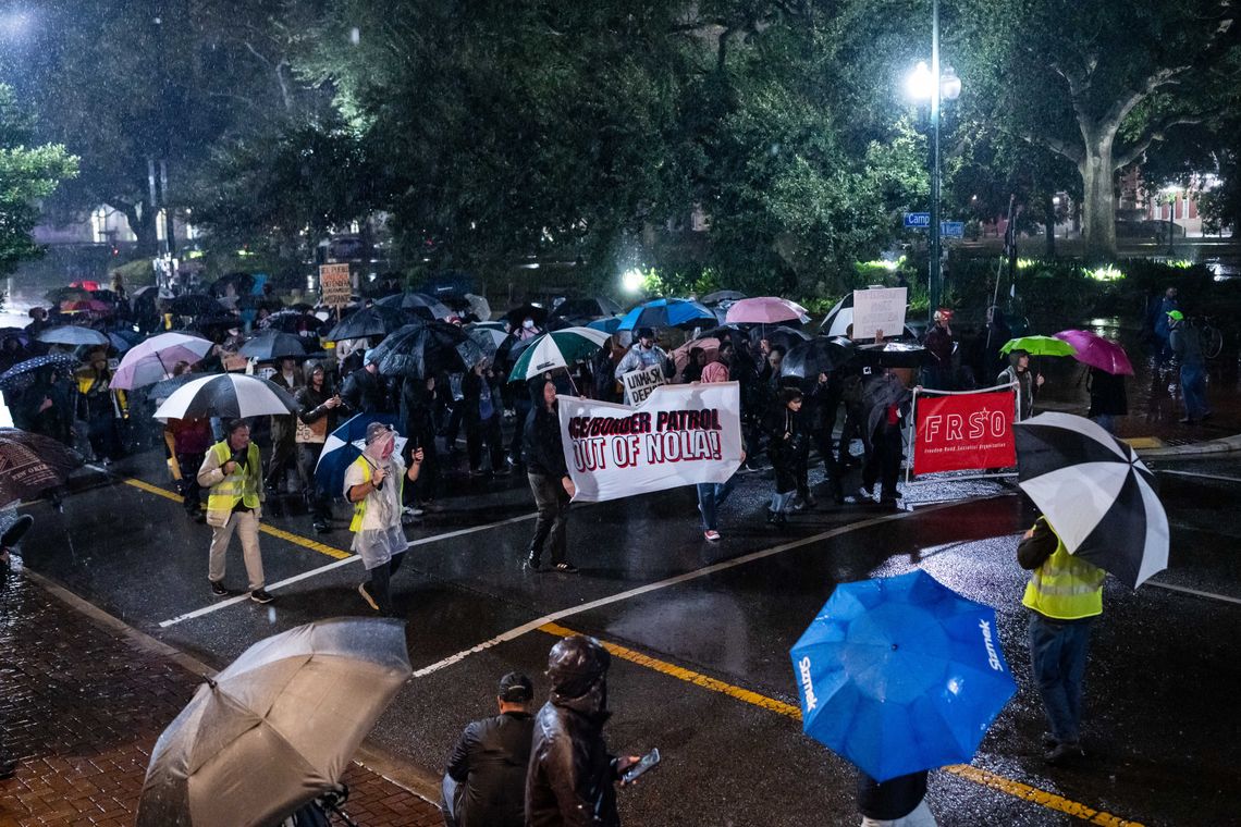 A group of protesters march in the street at night in the rain. People at the front of the group hold a banner that reads “ICE/Border Patrol out of NOLA.” 