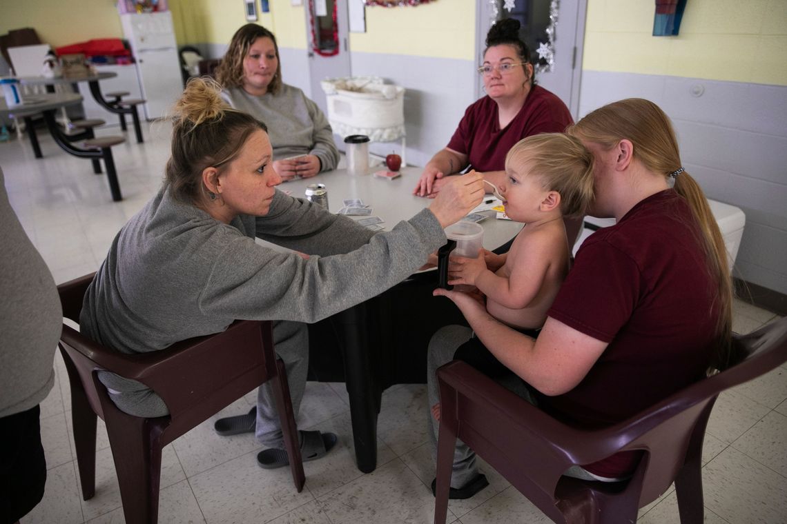 A photo shows a White woman with blond hair in a gray prison uniform feeding a blond baby held by another woman. They are sitting at a table with other women in a prison cafeteria.