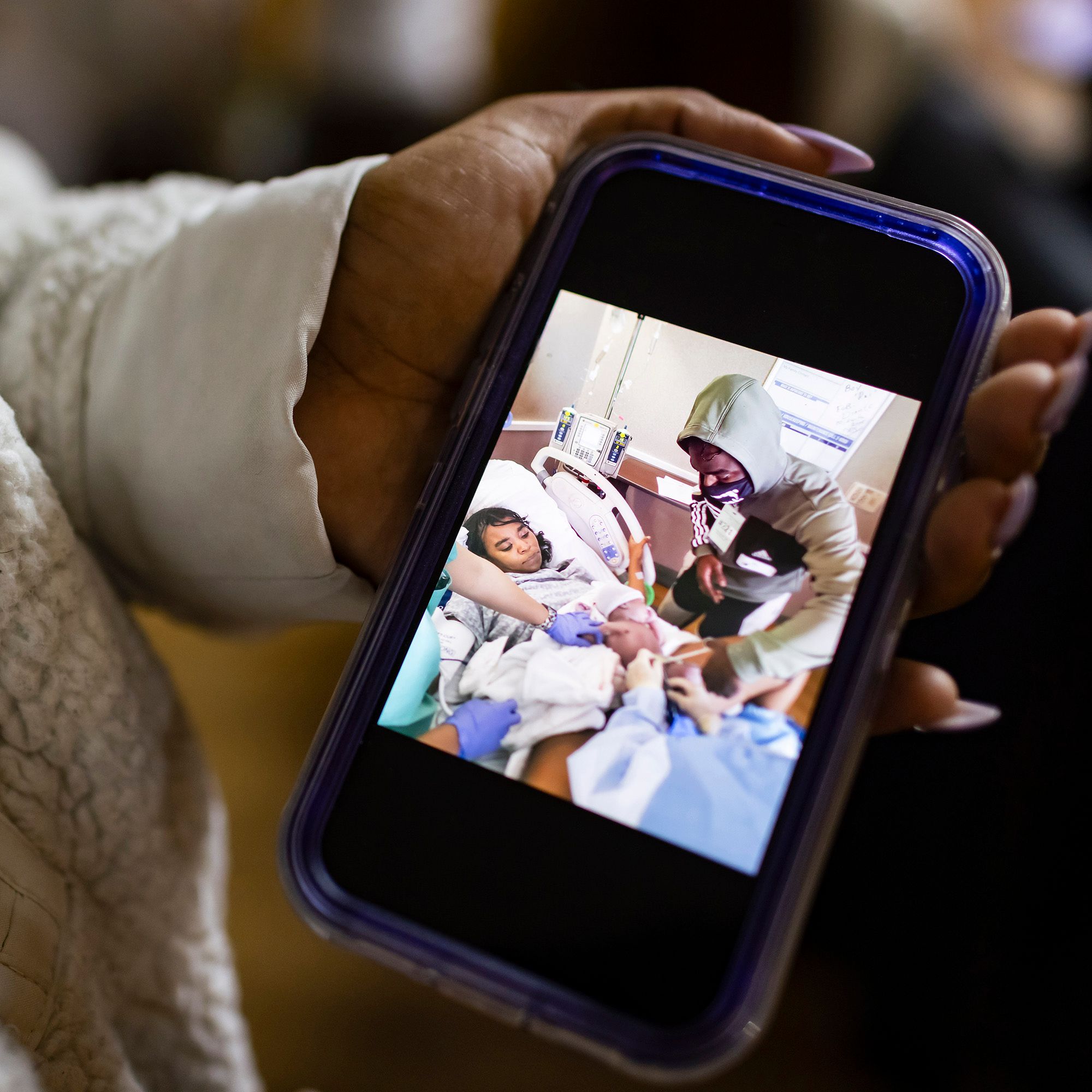 A photo shows a Black woman with purple nails and a white sweater holding her phone. The phone displays a picture of her husband, a Black man wearing a green hooded sweatshirt, cutting the umbilical cord of their baby after she gave birth. 