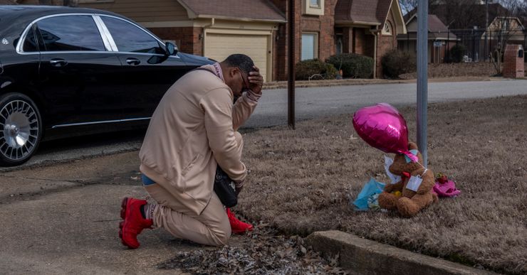 A Black man kneels at the side of a street, where a memorial is situated that has flowers, a teddy bear, and a pink ballon. 