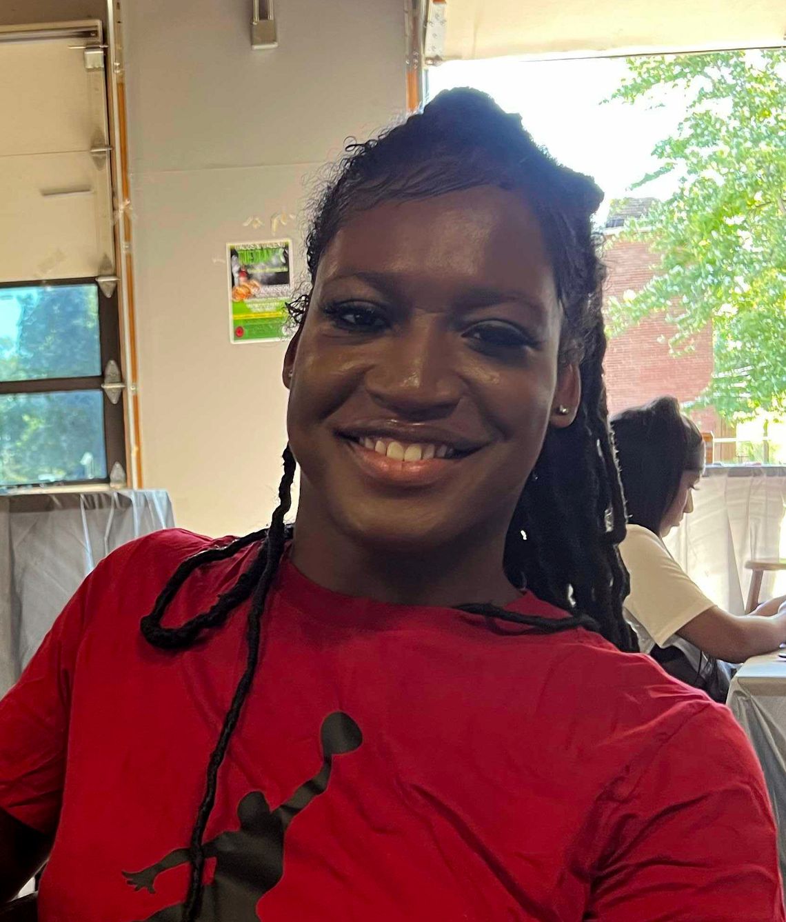 A Black transgender woman, wearing a red shirt, smiles for a photo.