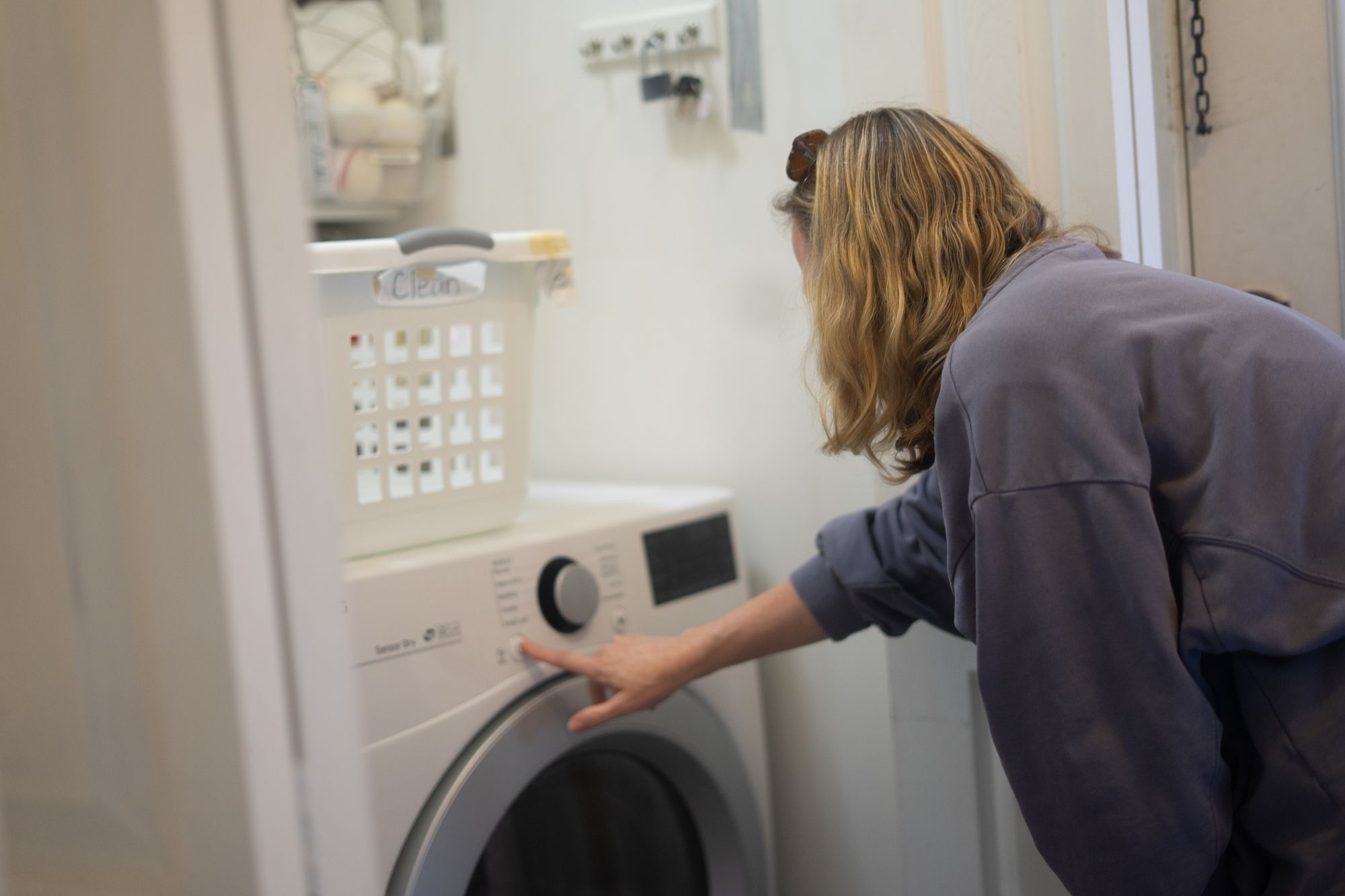 A woman with medium-length, light-brown hair and sunglasses on her head presses a button on a laundry machine. A white laundry basket sits on top of the machine.