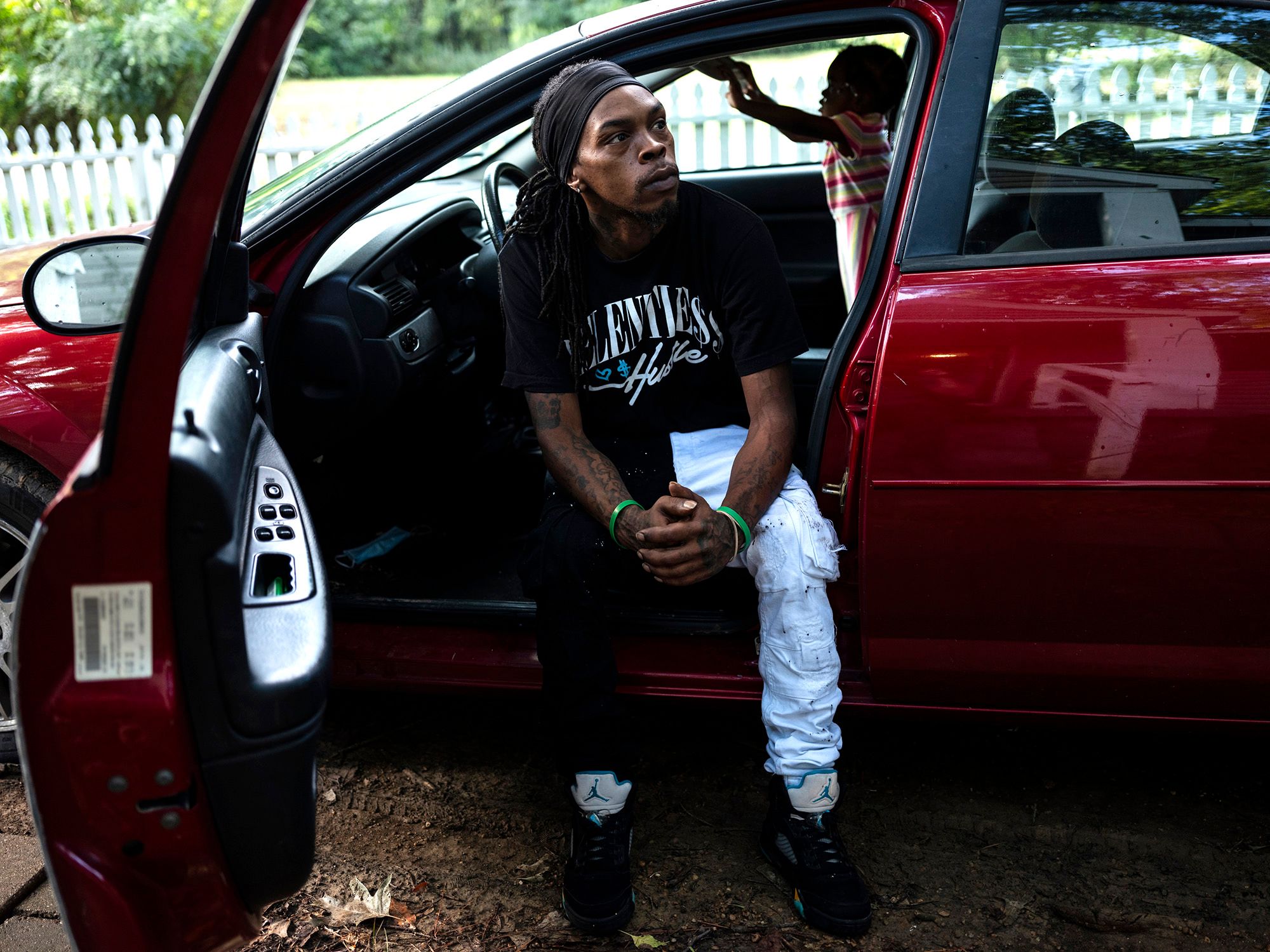 A Black man, wearing a black headband, black graphic T-shirt and black and white jeans, sits in the driver’s seat of a red car with the door opened. His young daughter plays inside the car in the background. 