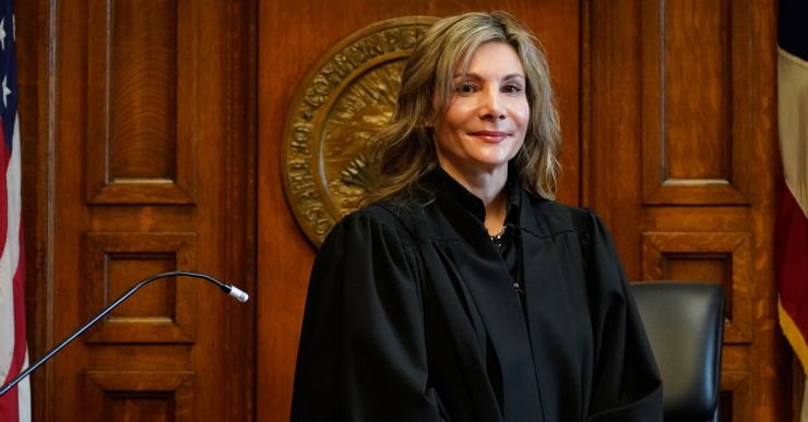 A White, blonde woman stands in a black robe behind a judge’s podium.