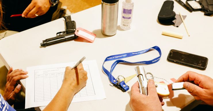 A close-up shot of three peoples' hands at a table. A medium-skin person is writing on a piece of paper. Also on the table, there is a black pistol with a red tag, a water container, hand sanitizer, a blue lanyard with the words "Bureau of Organized Crime," a phone ,and the hands of a light-skinned person unloading a silver pocket pistol. 