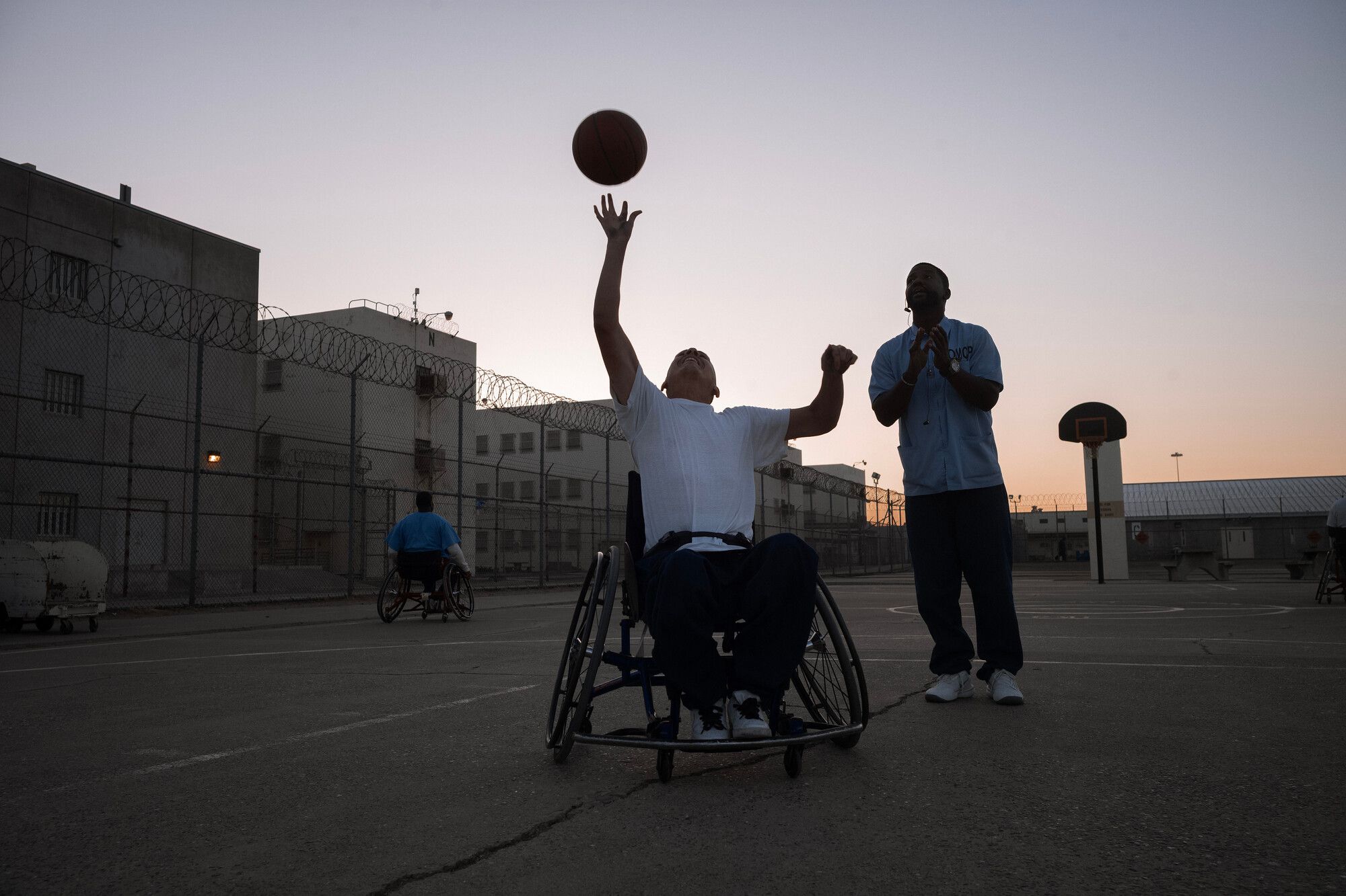 A photo shows a man in a wheelchair reaching up with the basketball in the air a few inches above his fingers. He is outdoors at dawn while another person stands nearby. 