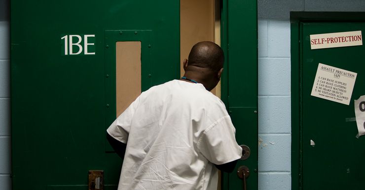 A Black medical ward assistant in a white uniform looks through an open door. 