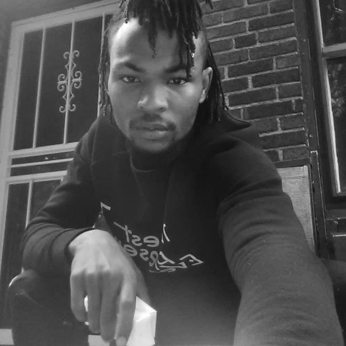 A black-and-white photo of a Black man with locs and a goatee. The man is looking into the camera while crouching down in front of a brick house. 
