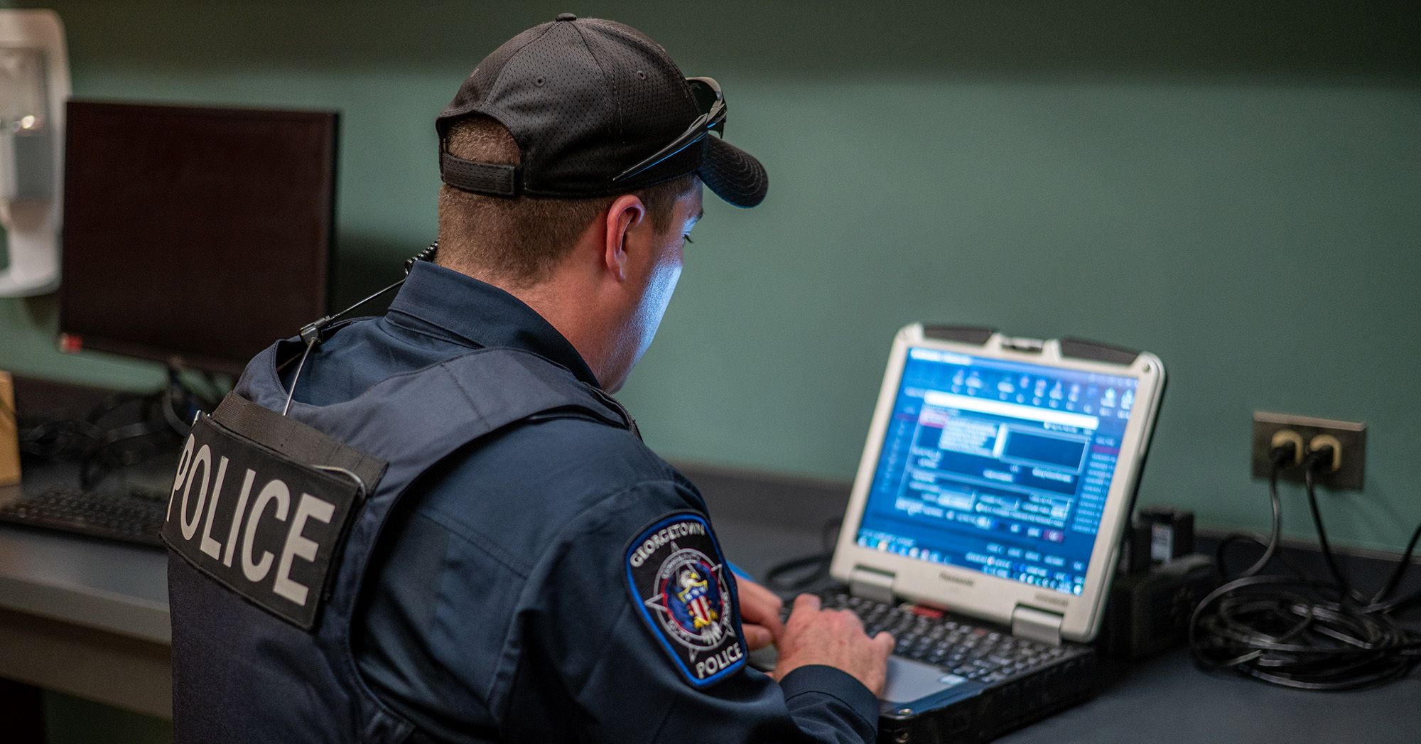 A police officer, wearing a cap, a vest and navy uniform, looks at a computer screen while sitting at desk. 