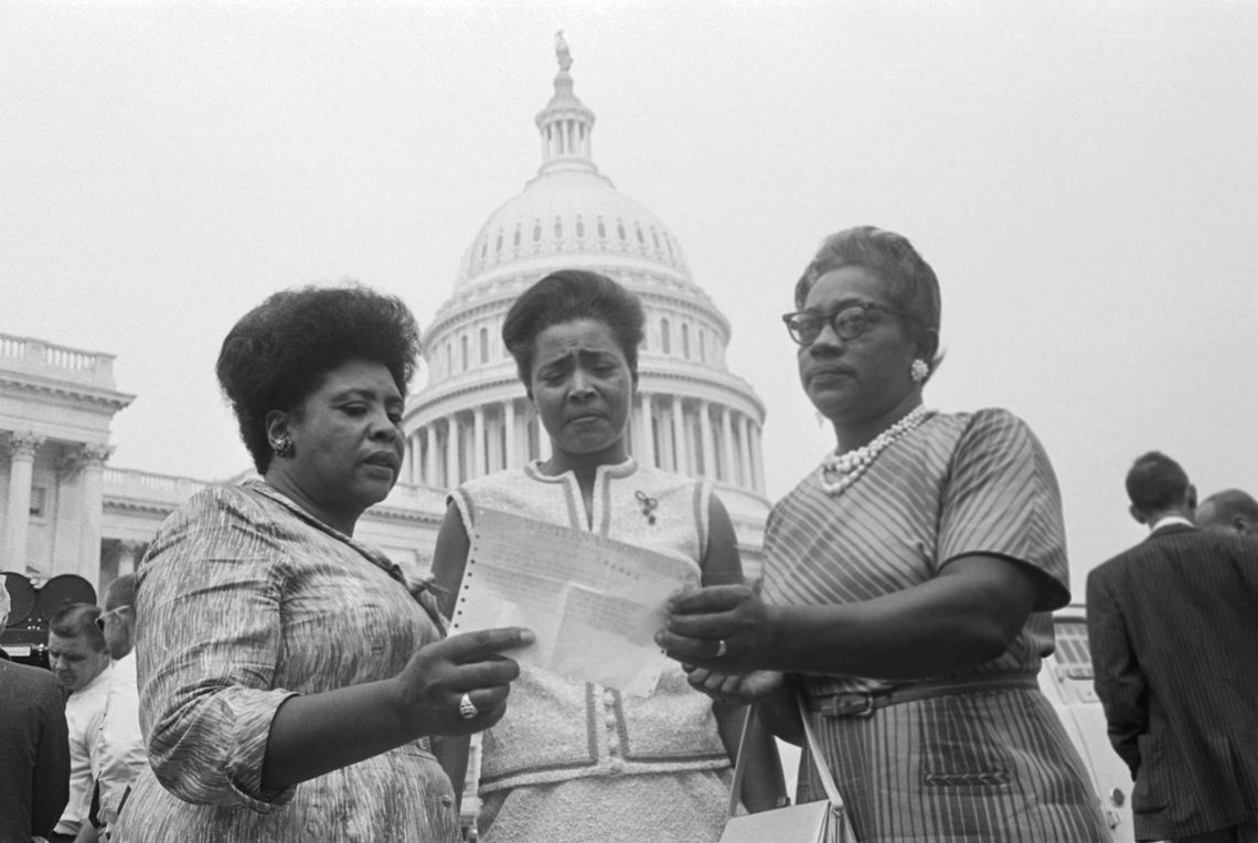 Three Black women with short hair stand in front of the U.S. Capitol, reading a piece of paper they’re holding. 