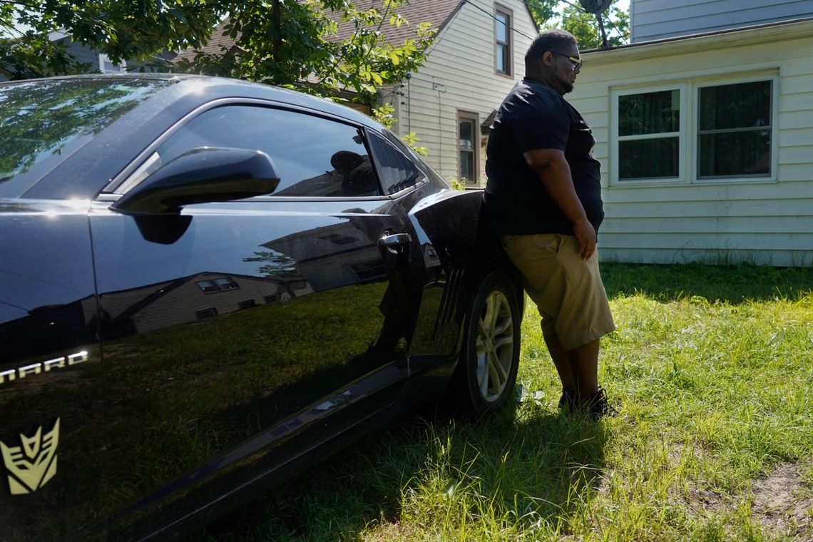 Henry Ray, a Black man, wears a black T-shirt, khaki shorts and sunglasses while leaning against his Camaro.