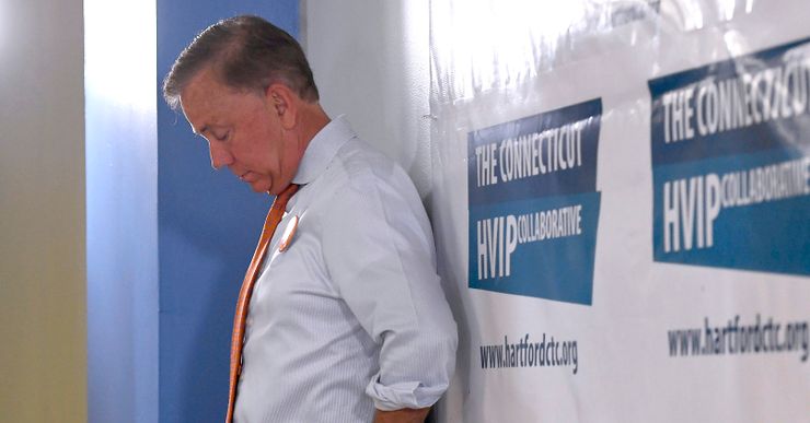 A White man is looking down with his hands on his hips, while leaning against a wall. He is dressed in a white button-up shirt with rolled-up sleeves, orange and black pants. 