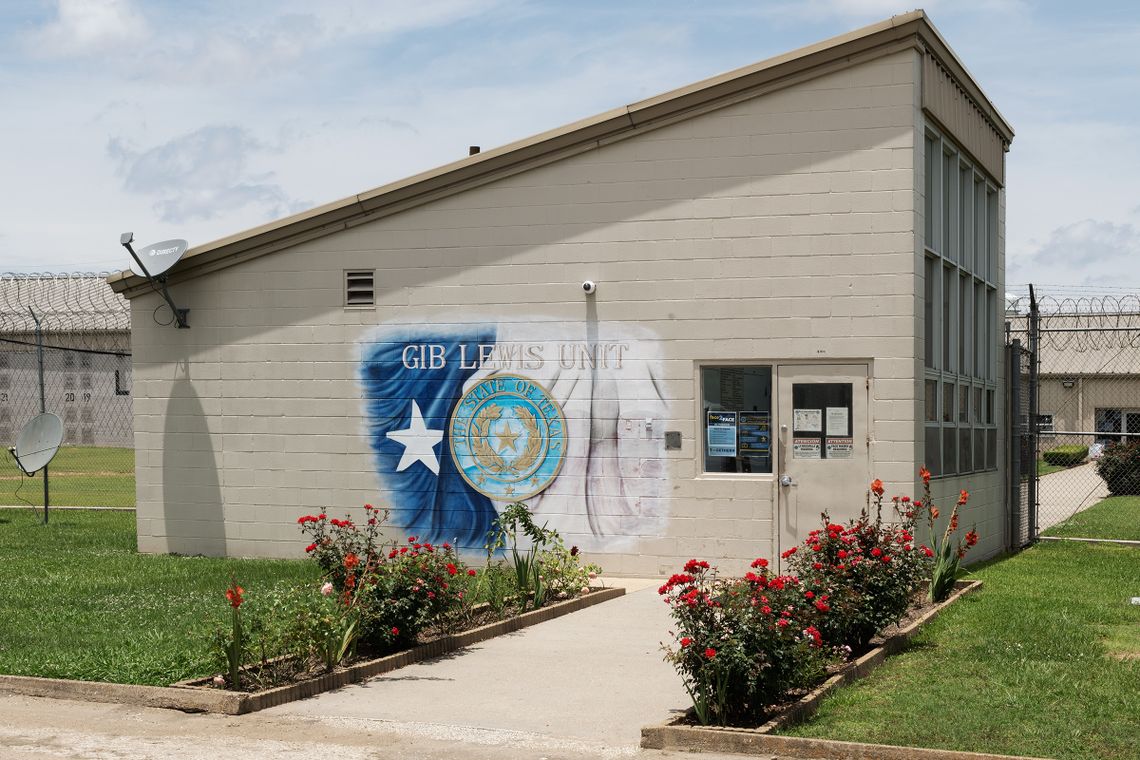 A small building on the campus of the Gib Lewis Unit stands in front of barbed wire. A painting showing the words "Gib Lewis Unit", part of the Texas flag with a white star on a blue background, and the seal of Texas are on the side of the building. 