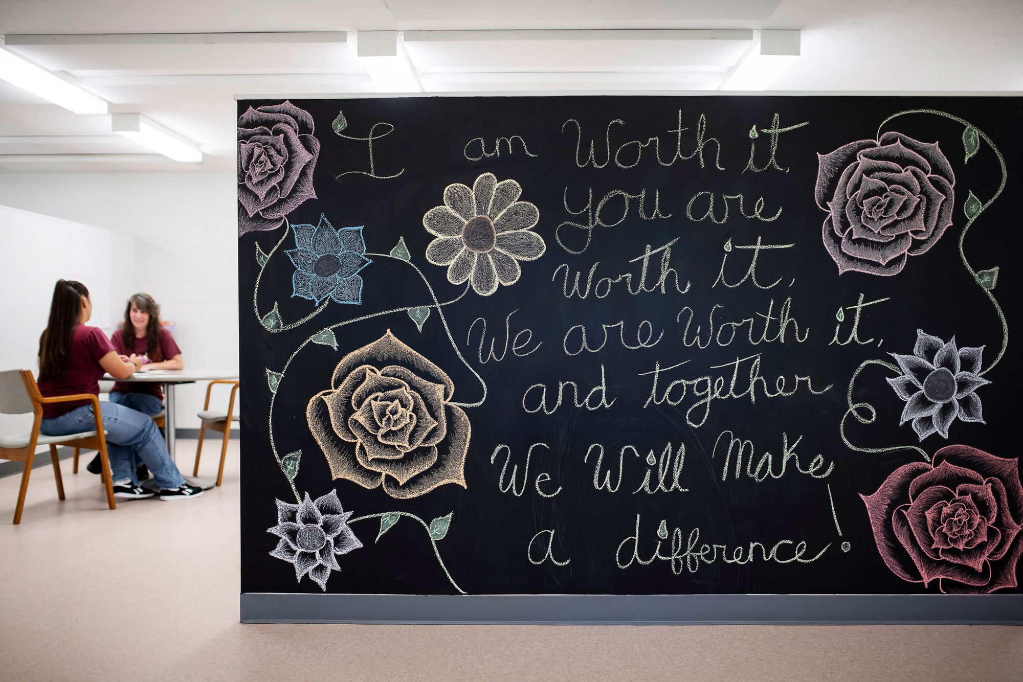 A room shows a black chalkboard towards the right side, reading “I am worth it; You are worth it; We are worth it, and together we will make a difference!” Two women wearing burgundy-colored t-shirts sit at a table on the left side in the background.