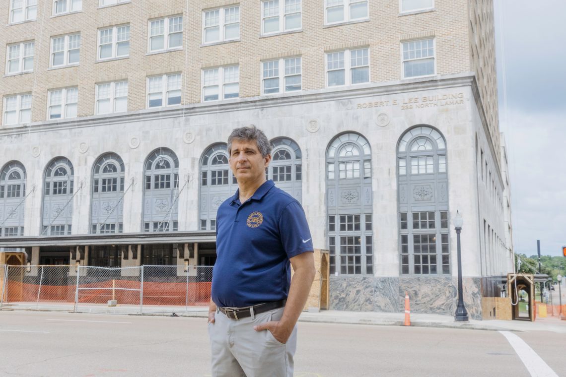 In a photograph, a White man with gray hair stands on the street in front of a concrete building with arched windows. The man is wearing a blue short-sleeved polo shirt with a logo. 