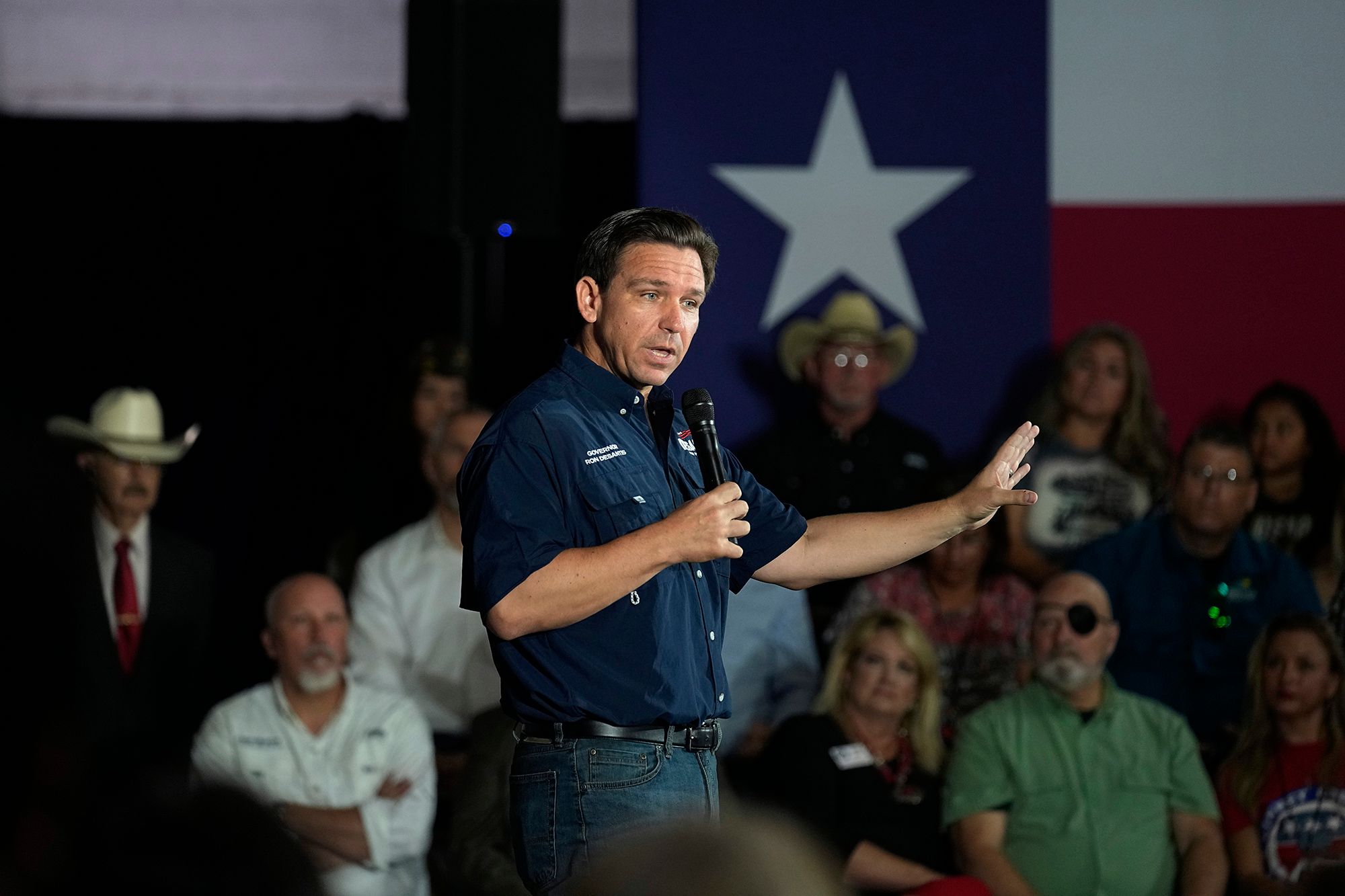 A White man, wearing a short-sleeved blue button up shirt and jeans, holds a microphone as he speaks to a crowd. Several people are seated in the background. A Texas flag hangs from the wall. 