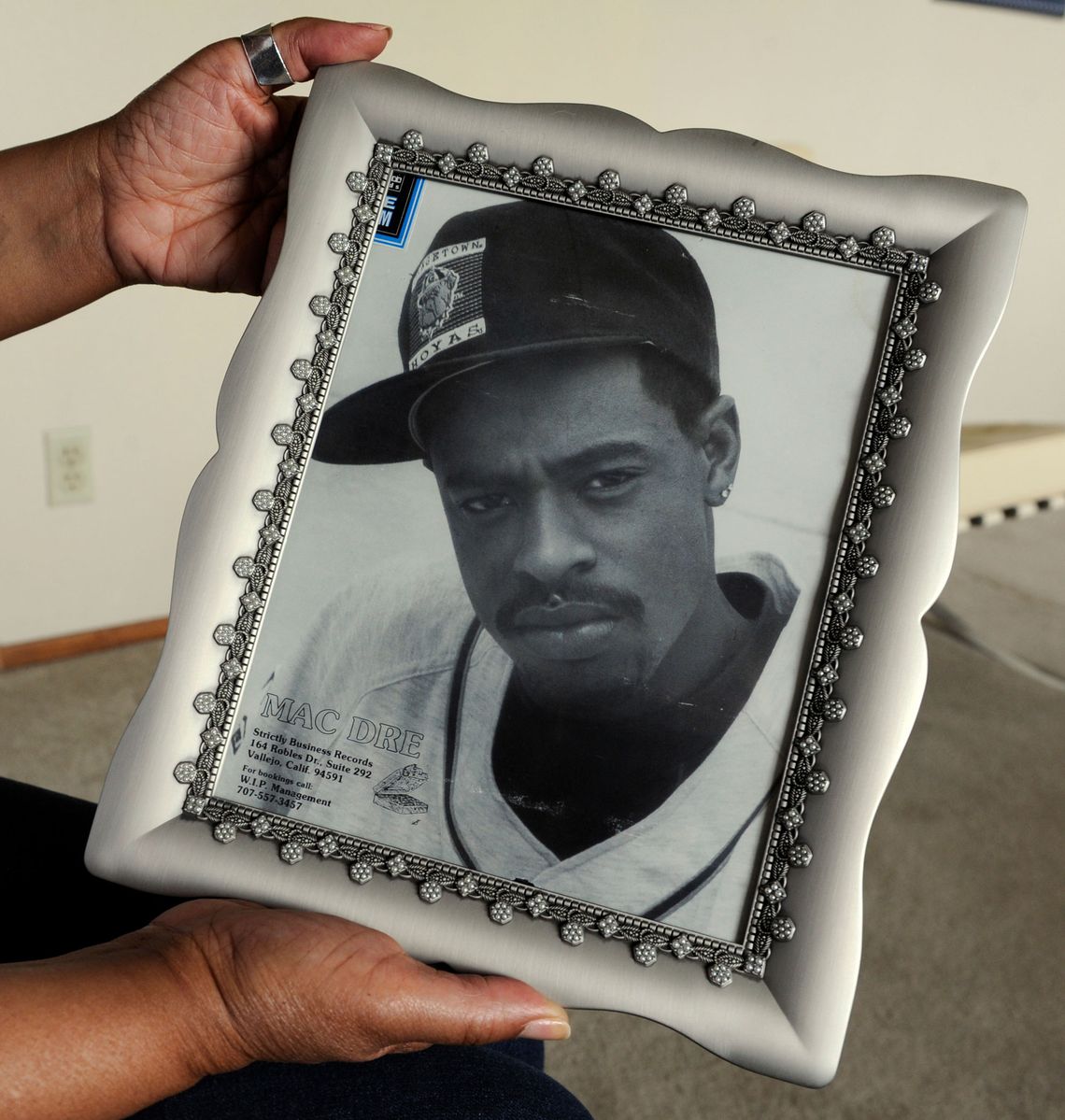 The hands of a Black woman hold the silver-colored framed black-and-white photo of her son, a young Black man wearing a dark-colored baseball cap with the logo of the Georgetown University Hoyas bulldog, a neatly trimmed goatee, a studded earring, and a light-colored baseball-style jersey. 