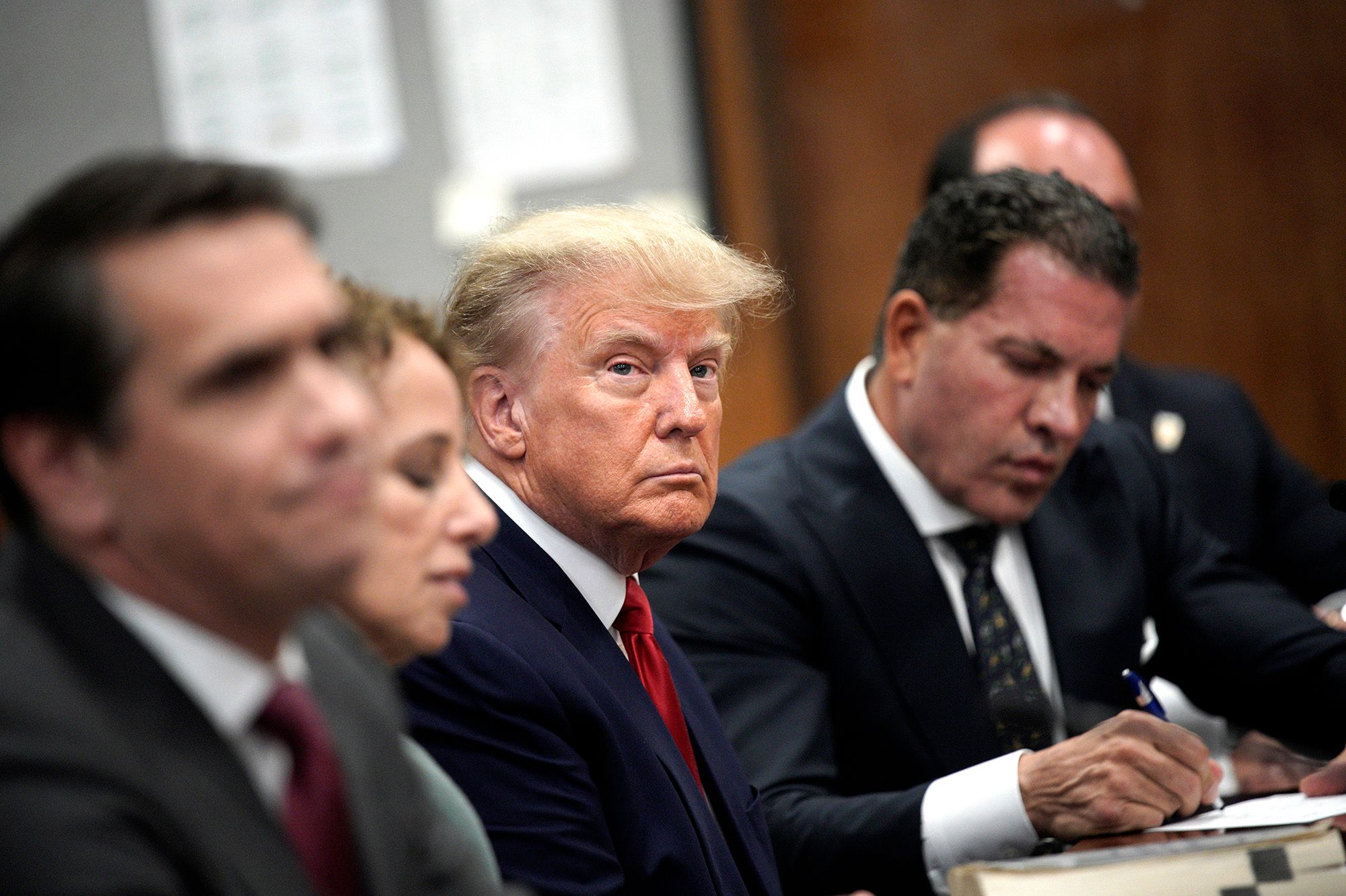 A White man, in a red tie and navy suit, sits in between his four attorneys in a courtroom.