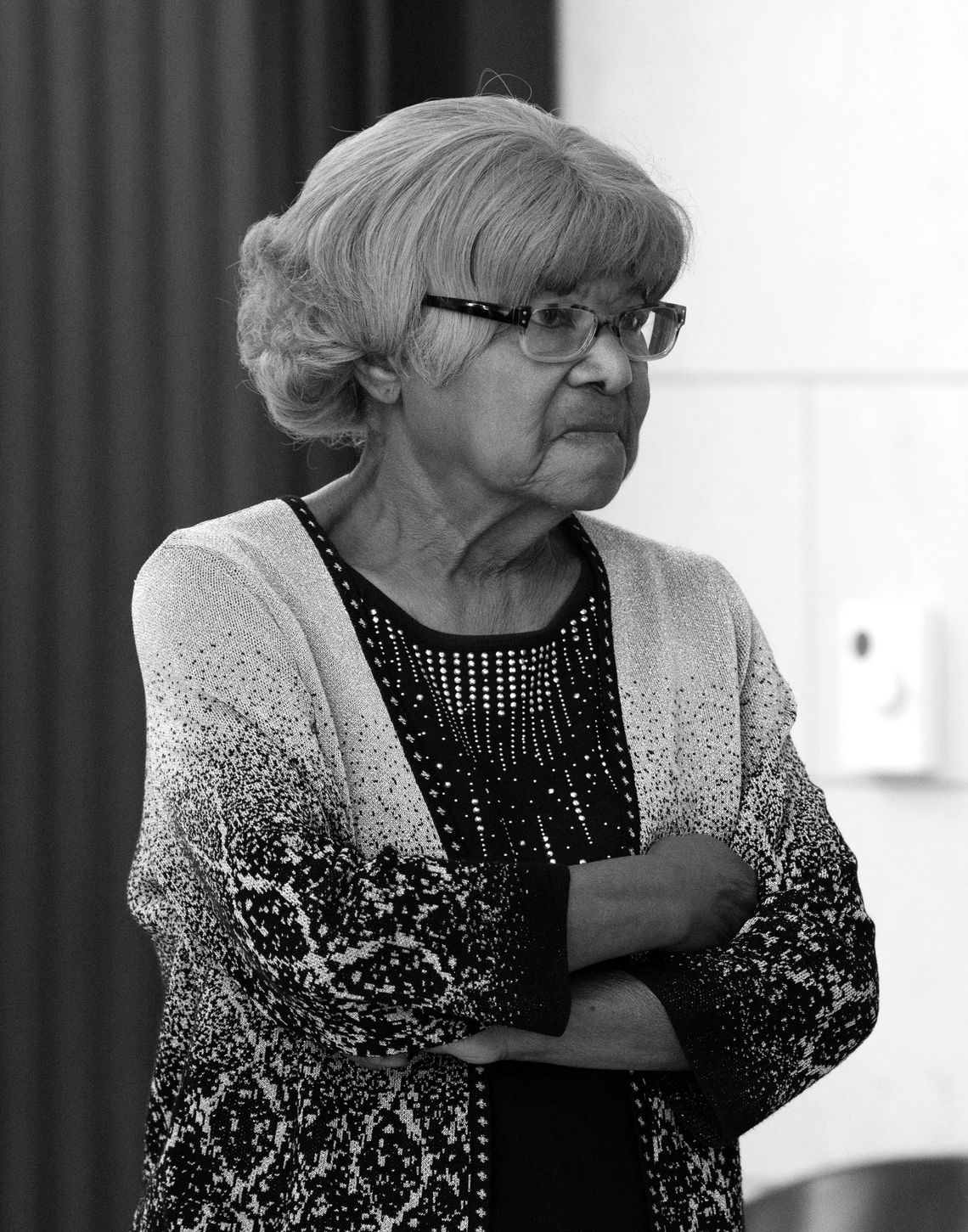 An older Black woman wearing glasses is standing with her arms crossed.
